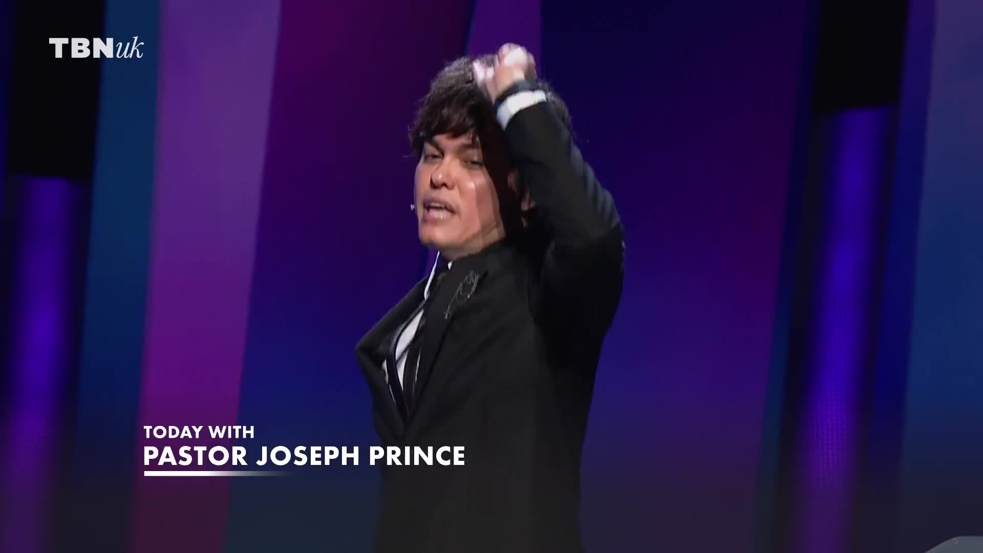Pastor Joseph Prince, on TBN UK, is gesturing with his right hand, running it through his dark hair. He's wearing a black suit and white shirt, speaking on a stage with a purple backdrop.
