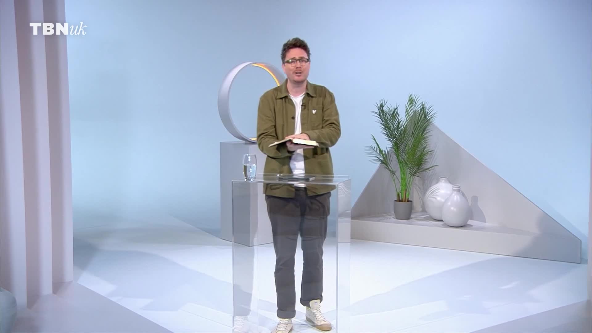 A man in a green jacket stands behind a clear podium, holding a book open in front of him. He's in a studio setting, with a modern, minimalist background.
