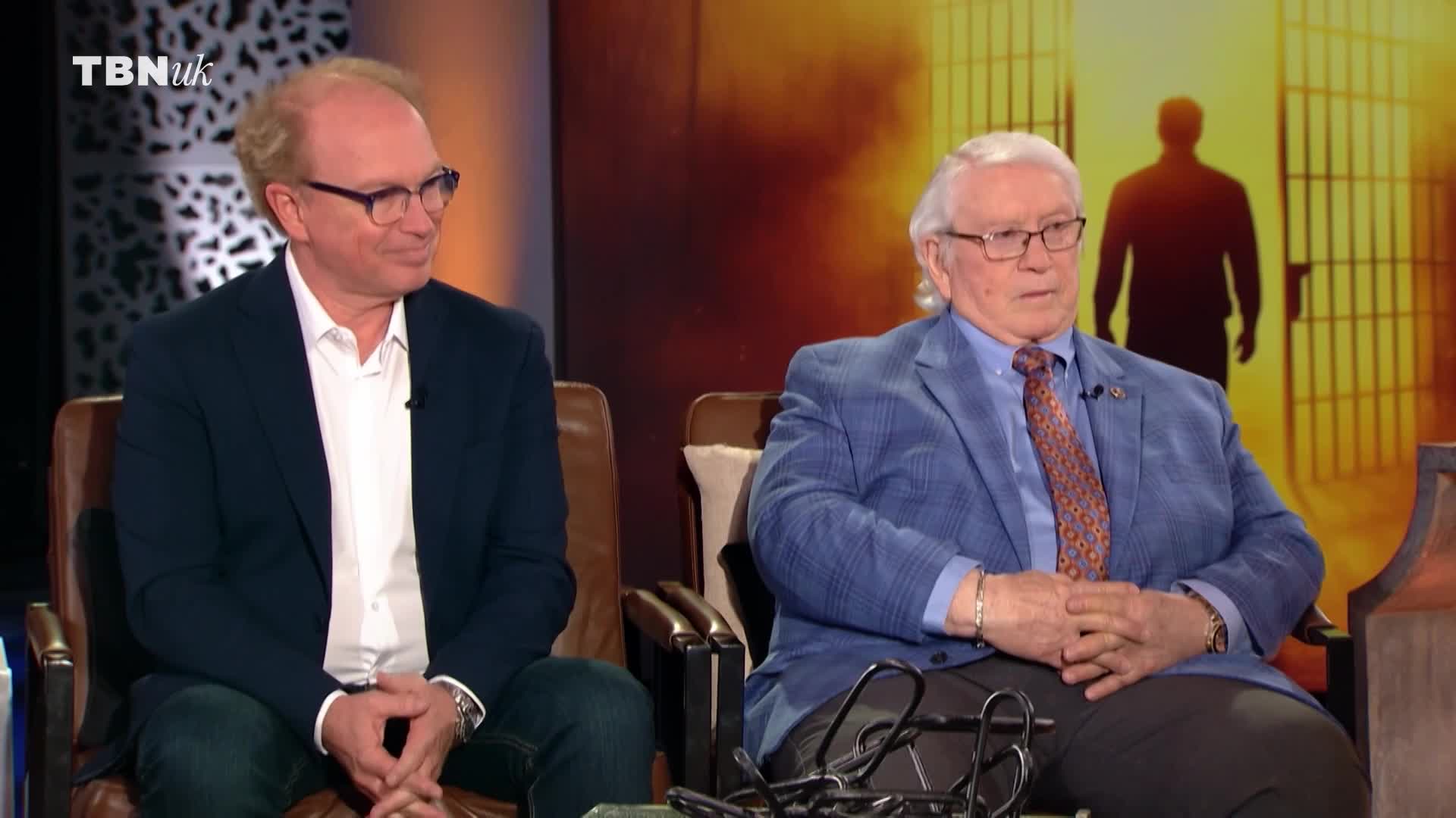 Two men in suits are seated in armchairs, facing forward. The backdrop on TBN UK shows a silhouette of a person walking out of a jail cell.
