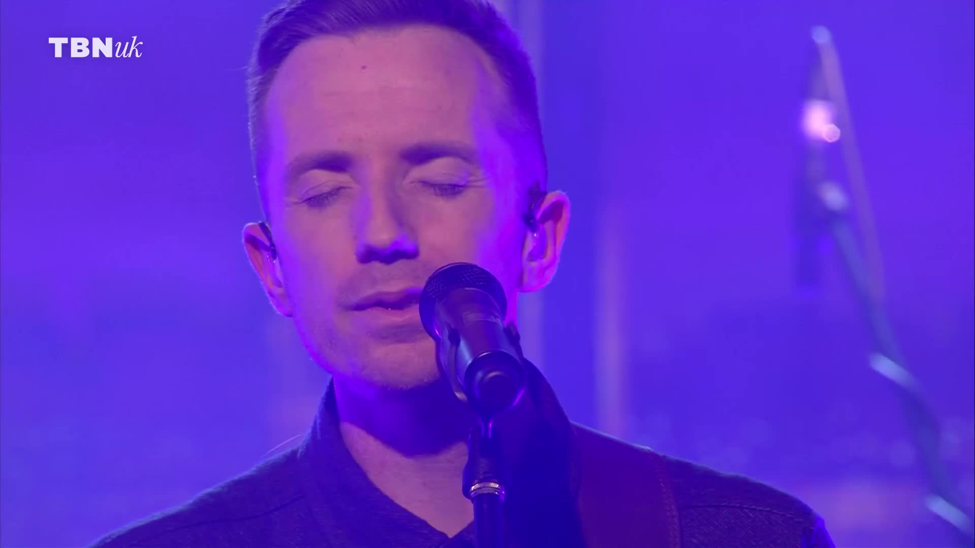 The man is singing, eyes closed, into a microphone.  He's on stage, bathed in purple light, and the TBN UK logo is visible in the corner.
