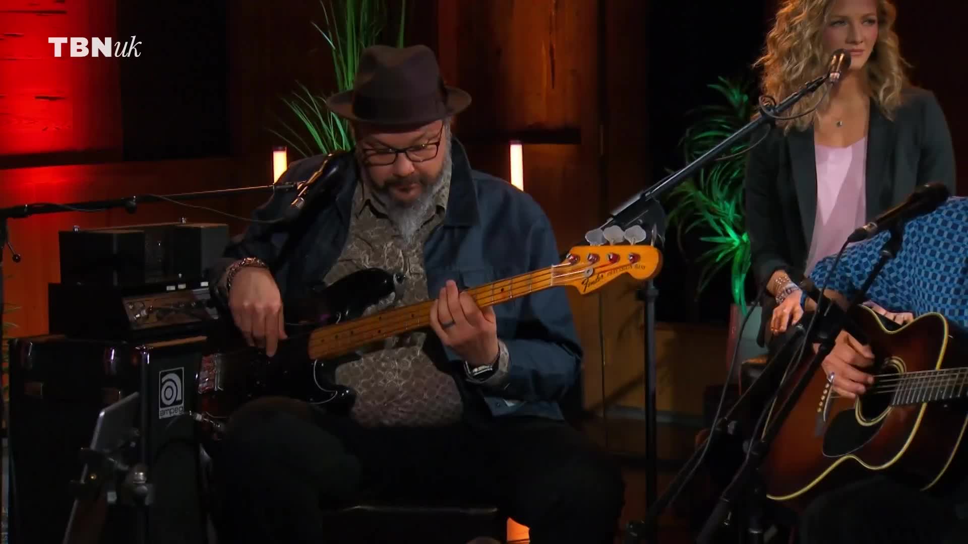 A bassist, wearing a fedora, plucks at the strings of his black bass guitar.  Behind him, a woman with blonde curls sings into a microphone, as the TBN UK logo sits in the corner.
