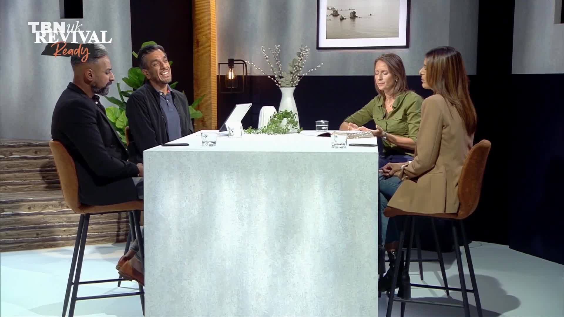 Four people sit at a table, engaged in what looks like a discussion, with one man leaning forward and smiling. The logo for TBN UK Revival Ready is visible in the upper left corner.
