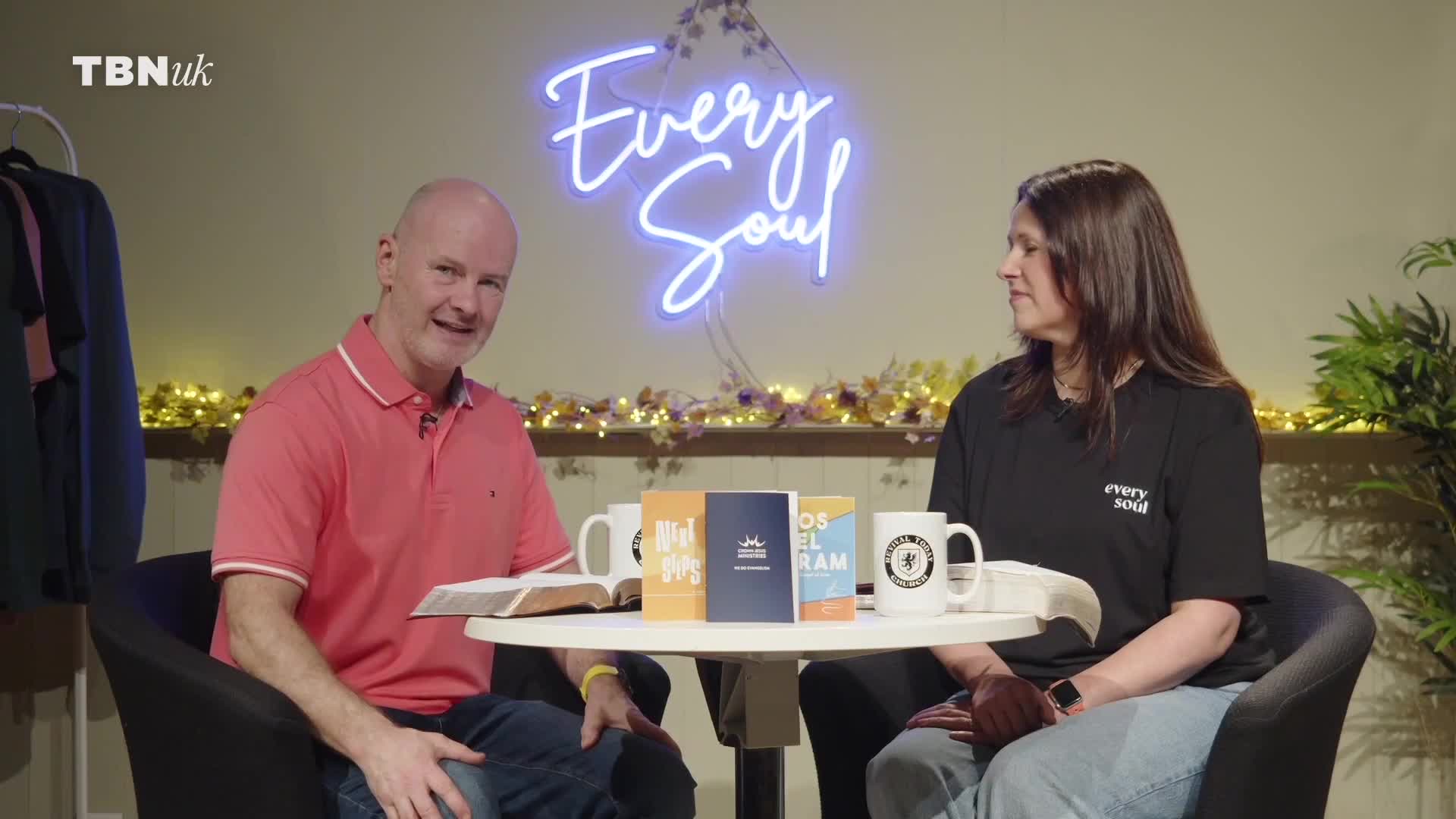 A man in a pink polo shirt and a woman in a black t-shirt sit at a table, facing the camera. Behind them, a neon sign reads "Every Soul," and the TBN UK logo is visible in the corner.
