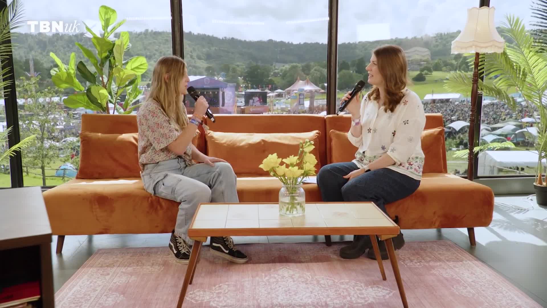 Two women, each holding a microphone, sit on a burnt orange couch, chatting. Behind them, a large window offers a view of a crowd gathered at a festival in the United Kingdom, likely for TBN UK.
