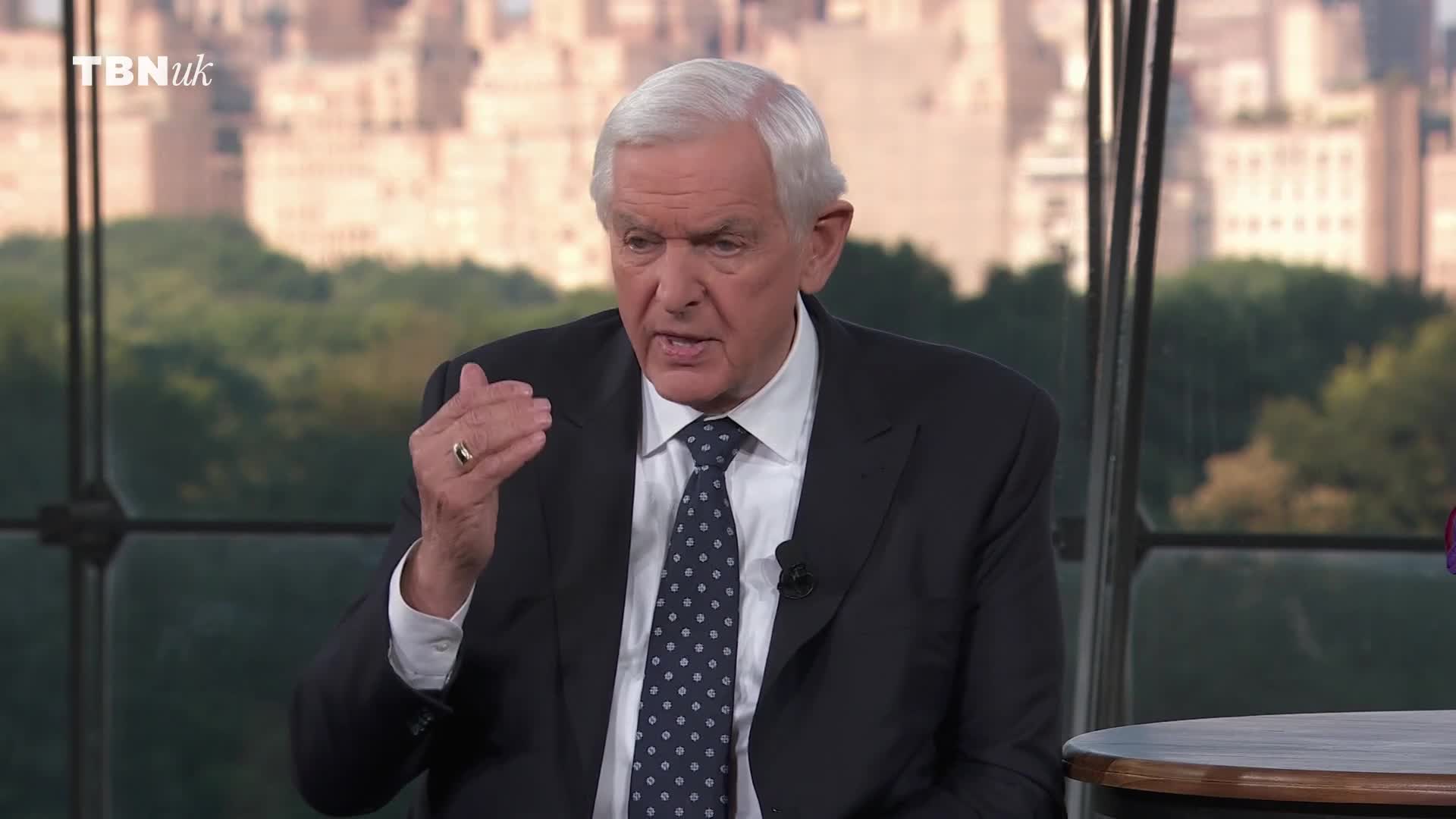 The man in the suit gestures with his right hand as he speaks. A ring glints on his finger, and the backdrop behind him suggests a city.
