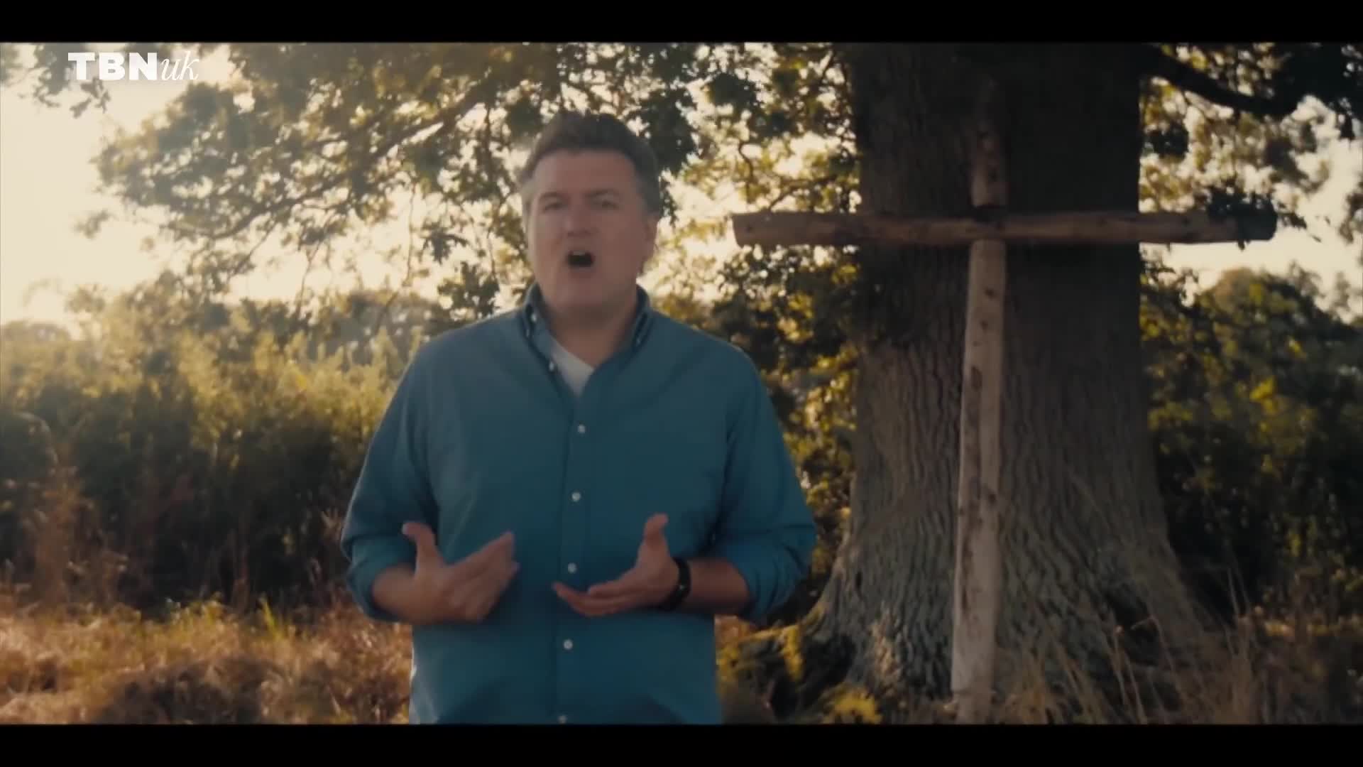 A man in a blue shirt gestures emphatically in front of a rustic wooden cross. The TBN UK logo is visible in the corner, and the scene is set outdoors, with a large tree in the background.
