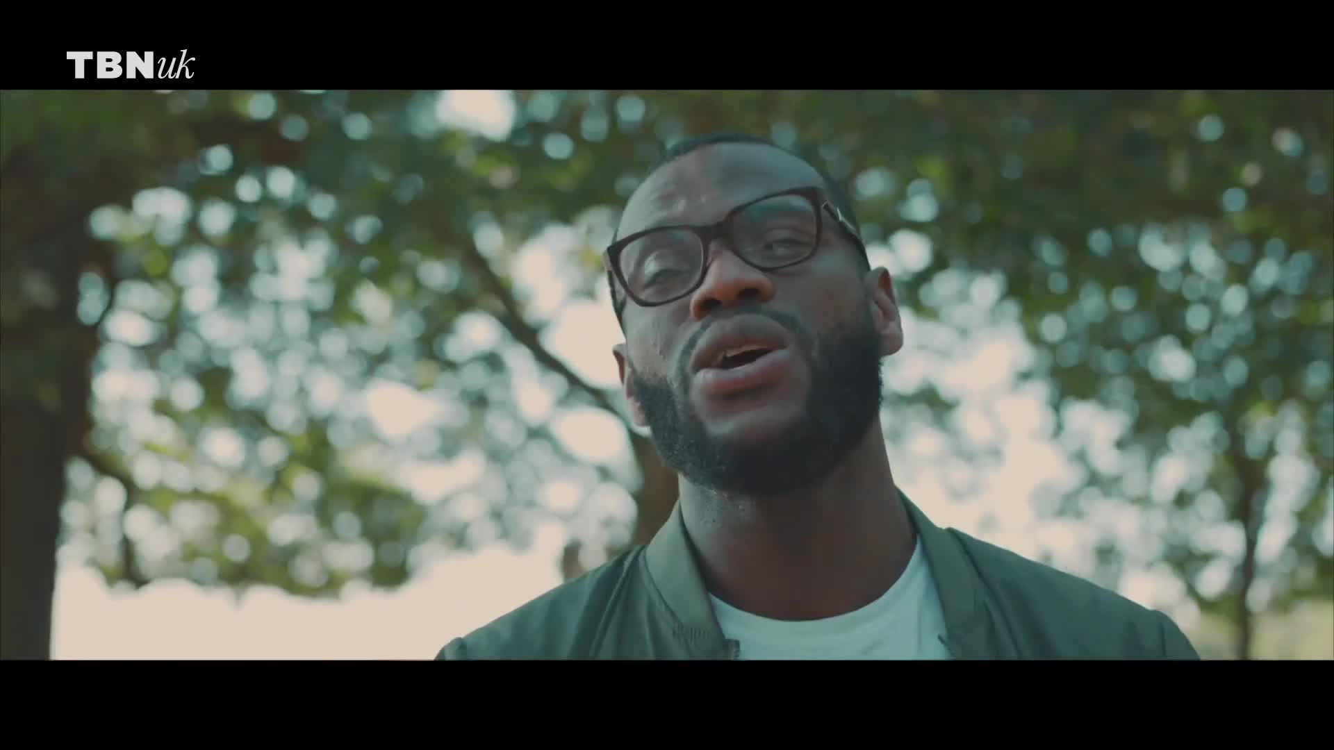 A man with glasses and a beard is speaking, his mouth moving as he looks toward the camera. Behind him, a soft focus of green leaves and branches suggests an outdoor setting, perhaps somewhere in the United Kingdom.
