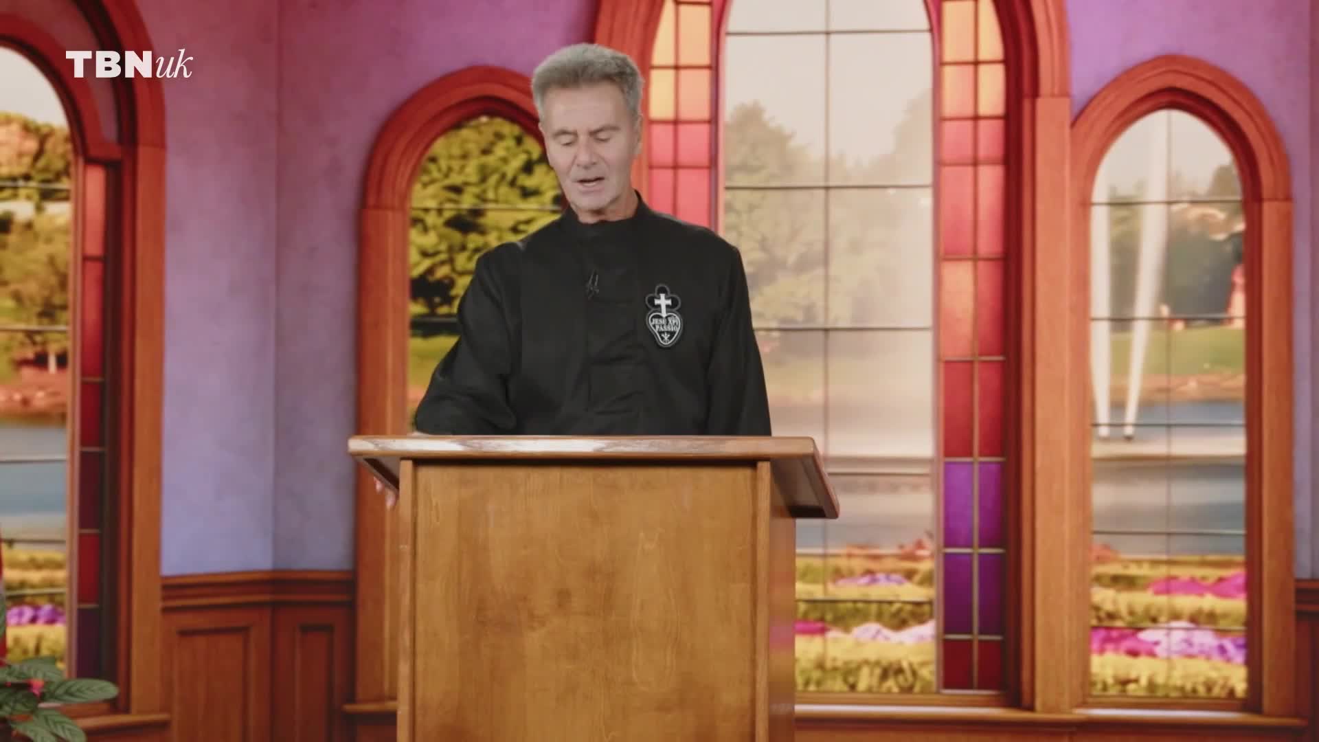 A man in a black jacket stands behind a wooden podium, speaking. Behind him, large arched windows offer views of a garden and what looks like a fountain, all bathed in soft light.
A man in a black jacket stands behind a wooden podium, speaking. Behind him, large arched windows offer views of a garden and what looks like a fountain, all bathed in soft light.