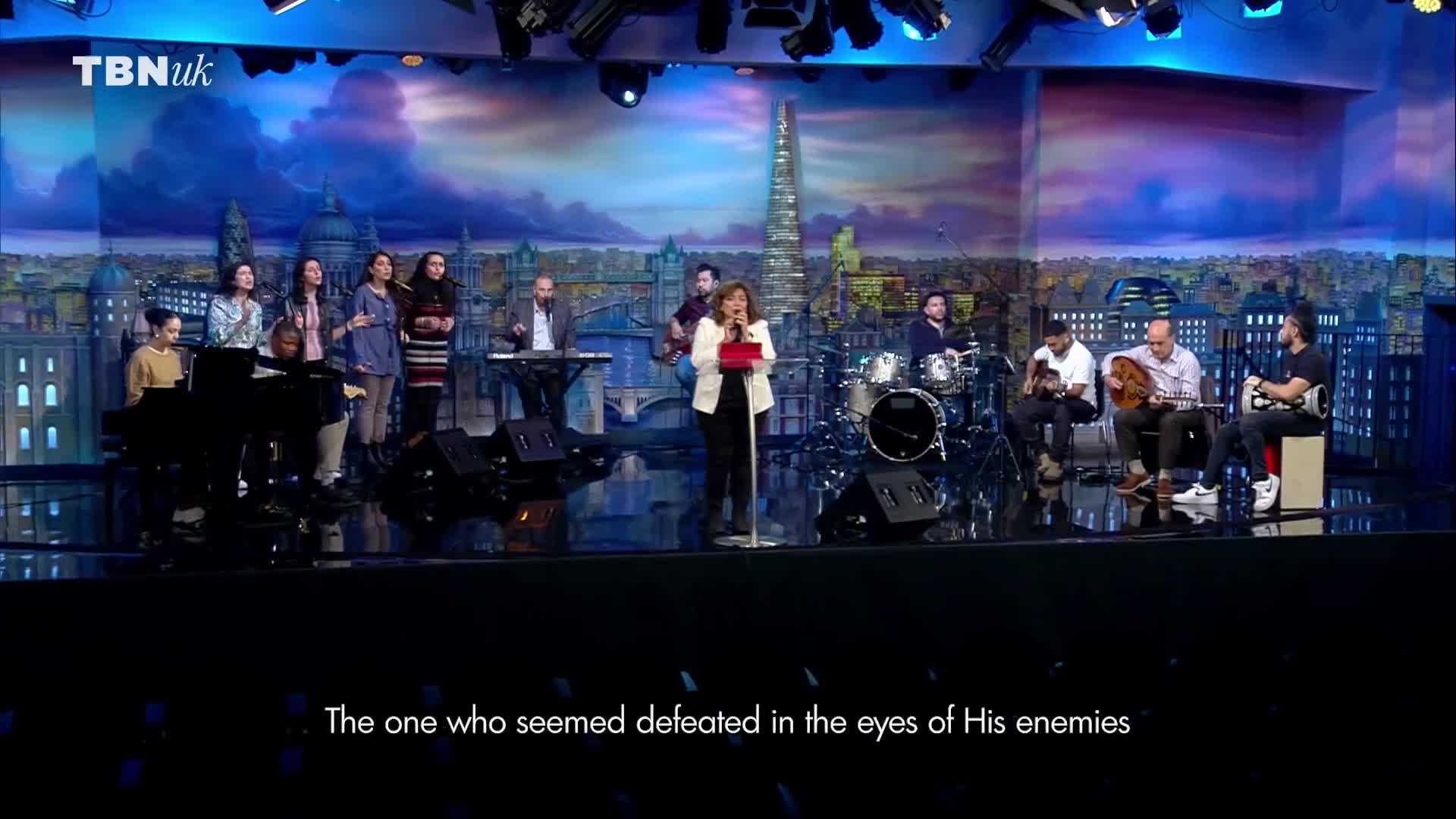 A woman in a blazer stands center stage, singing into a microphone, backed by a band and a choir. The backdrop depicts a nighttime cityscape of London, likely for a broadcast by TBN UK.
A woman in a blazer stands center stage, singing into a microphone, backed by a band and a choir. The backdrop depicts a nighttime cityscape of London, likely for a broadcast by TBN UK.