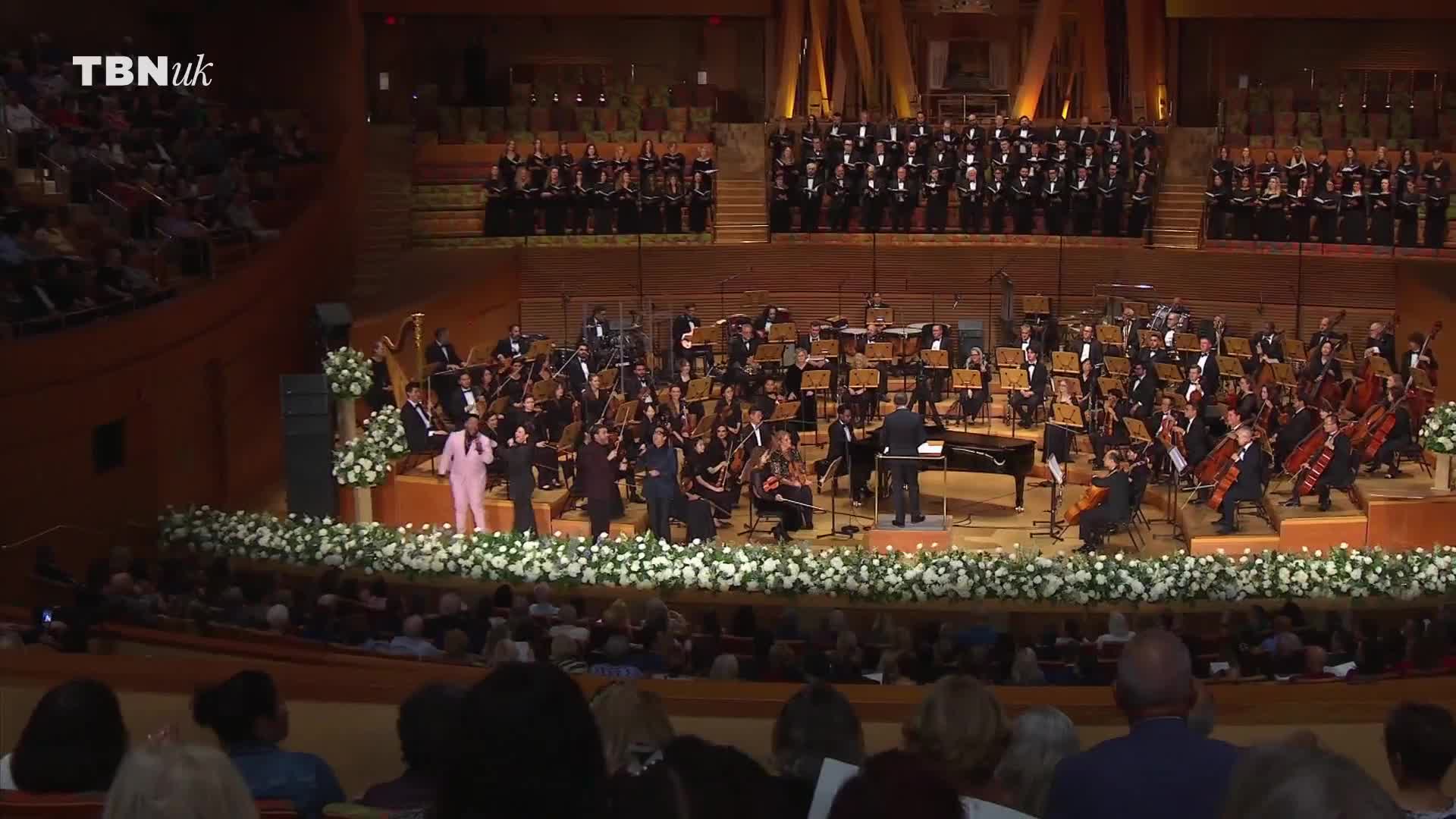 On stage, a group of singers in vibrant attire perform in front of an orchestra and a large choir. The conductor directs the musicians as the performance unfolds, captured by TBN UK.
On stage, a group of singers in vibrant attire perform in front of an orchestra and a large choir. The conductor directs the musicians as the performance unfolds, captured by TBN UK.