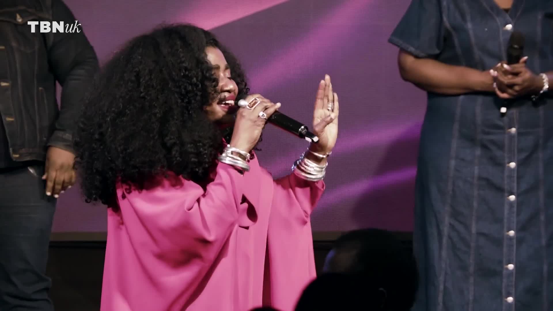 On the TBN UK stage, the woman in the pink dress sings into a microphone, her hand raised in a gesture. Her curly hair bounces as she moves, and she's surrounded by others.
On the TBN UK stage, the woman in the pink dress sings into a microphone, her hand raised in a gesture. Her curly hair bounces as she moves, and she's surrounded by others.