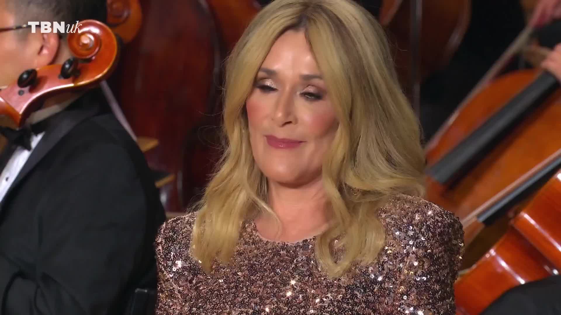 A woman in a sparkly dress looks down, a slight smile on her face. Behind her, a string orchestra is visible, and the TBN UK logo is in the corner.
A woman in a sparkly dress looks down, a slight smile on her face. Behind her, a string orchestra is visible, and the TBN UK logo is in the corner.