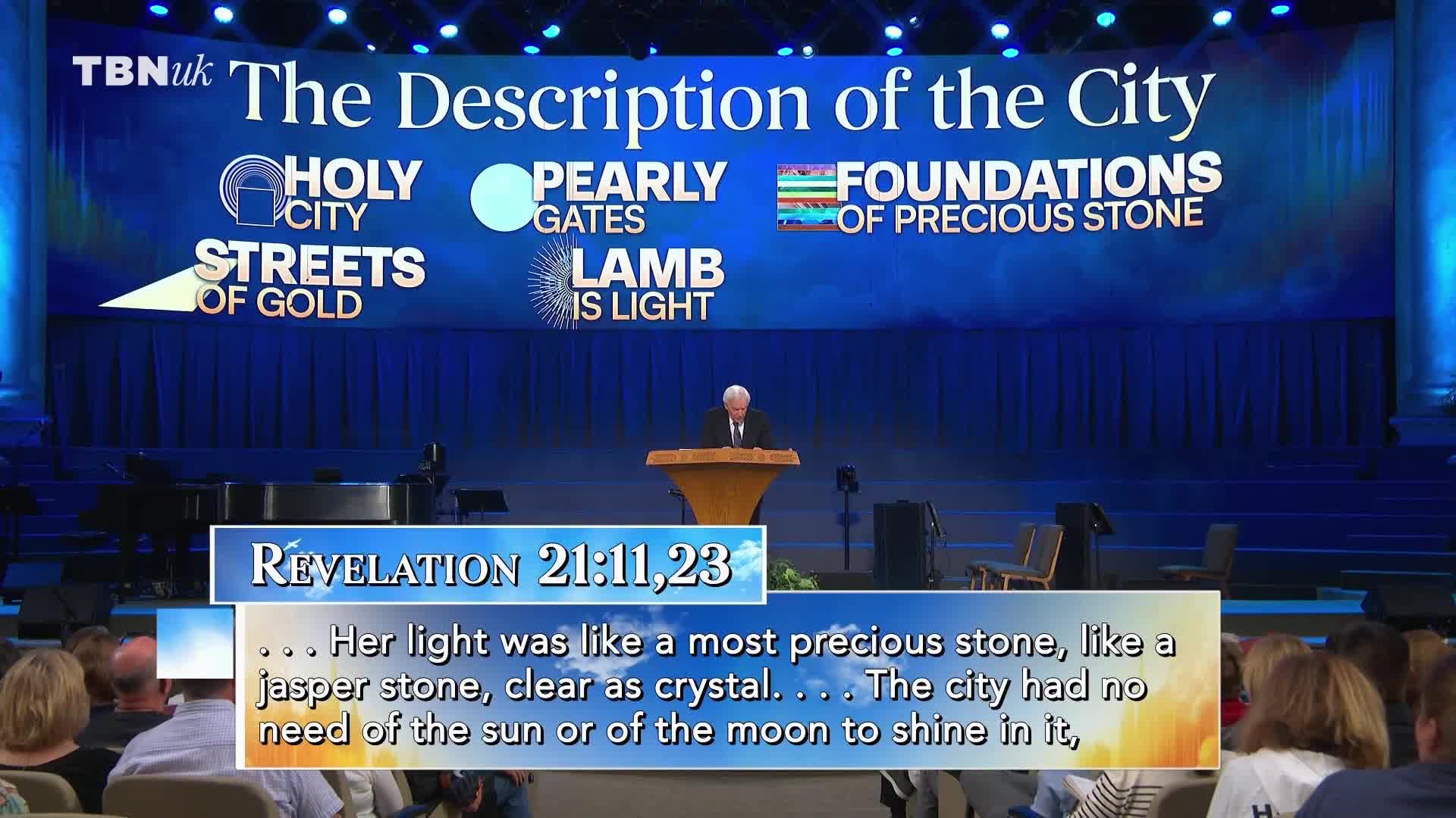 A man in a suit stands at a podium, reading from a text on a stage with a blue backdrop. Above him, a screen displays the TBN UK logo and the words "The Description of the City."
A man in a suit stands at a podium, reading from a text on a stage with a blue backdrop. Above him, a screen displays the TBN UK logo and the words "The Description of the City."