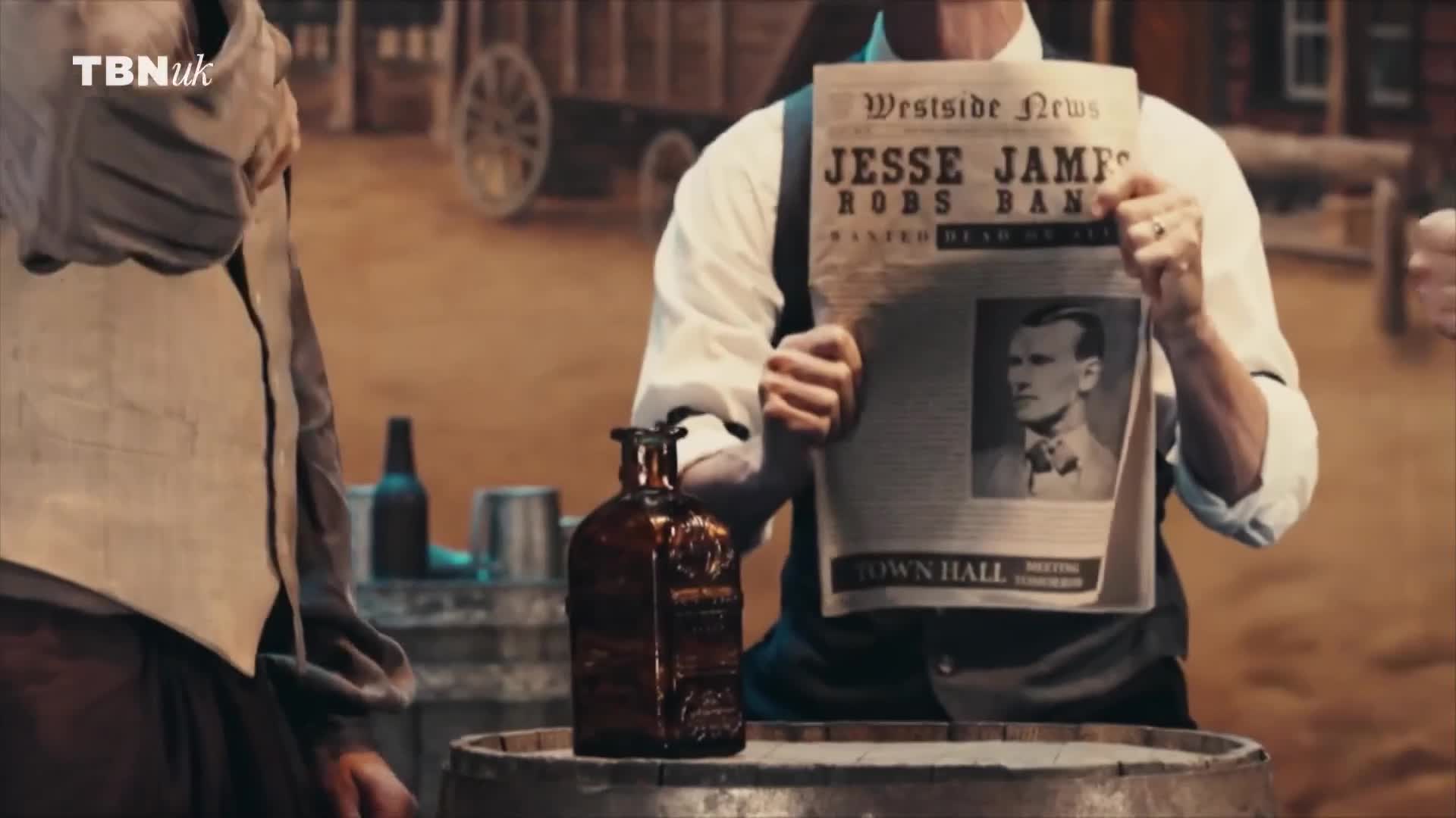 A man in a white shirt and suspenders holds up a newspaper with a photo of Jesse James, the headline reading "Jesse James Robs Bank." An amber bottle sits on a barrel in front of him, and the logo for TBN UK is in the top left corner.
A man in a white shirt and suspenders holds up a newspaper with a photo of Jesse James, the headline reading "Jesse James Robs Bank." An amber bottle sits on a barrel in front of him, and the logo for TBN UK is in the top left corner.