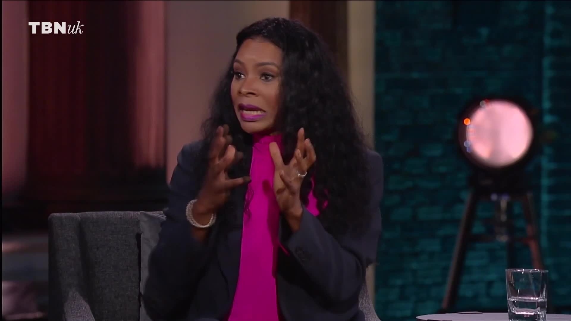 The woman, wearing a dark blazer and a bright pink top, gestures emphatically with both hands. She's sitting on a set for TBN UK, her expression intense as she speaks.
The woman, wearing a dark blazer and a bright pink top, gestures emphatically with both hands. She's sitting on a set for TBN UK, her expression intense as she speaks.