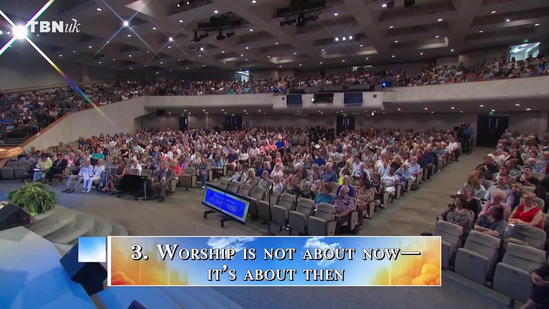 A large crowd is seated in rows, facing a stage. The TBN UK logo is visible, and a screen displays a message about worship.
A large crowd is seated in rows, facing a stage. The TBN UK logo is visible, and a screen displays a message about worship.