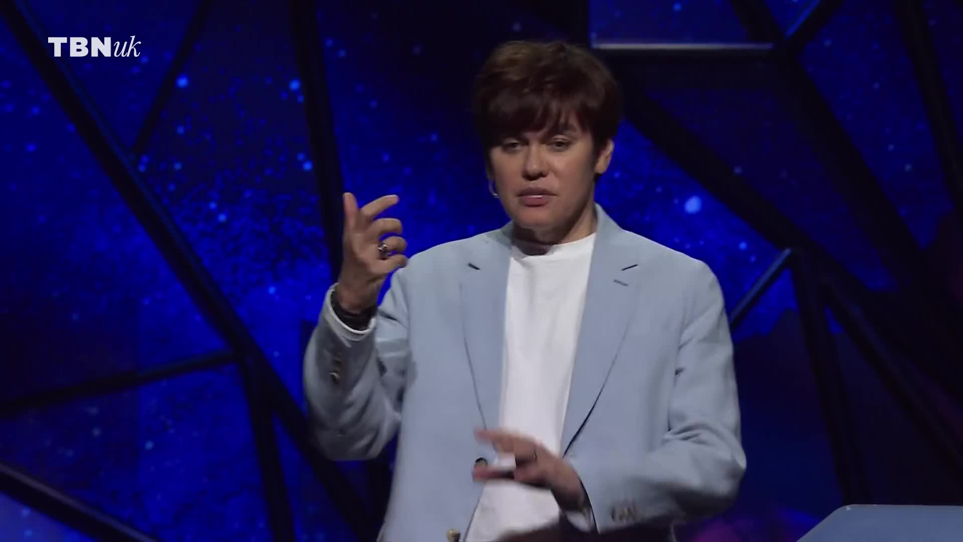 A man in a light blue jacket gestures with his hands, speaking on a stage in the United Kingdom for TBN UK. He's wearing a white t-shirt and a ring on his finger.
A man in a light blue jacket gestures with his hands, speaking on a stage in the United Kingdom for TBN UK. He's wearing a white t-shirt and a ring on his finger.