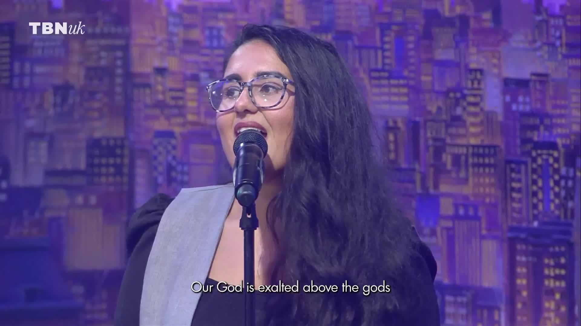 A woman with glasses sings into a microphone, her mouth open. The stage backdrop is a stylized cityscape, and the TBN UK logo is visible in the corner.
A woman with glasses sings into a microphone, her mouth open. The stage backdrop is a stylized cityscape, and the TBN UK logo is visible in the corner.