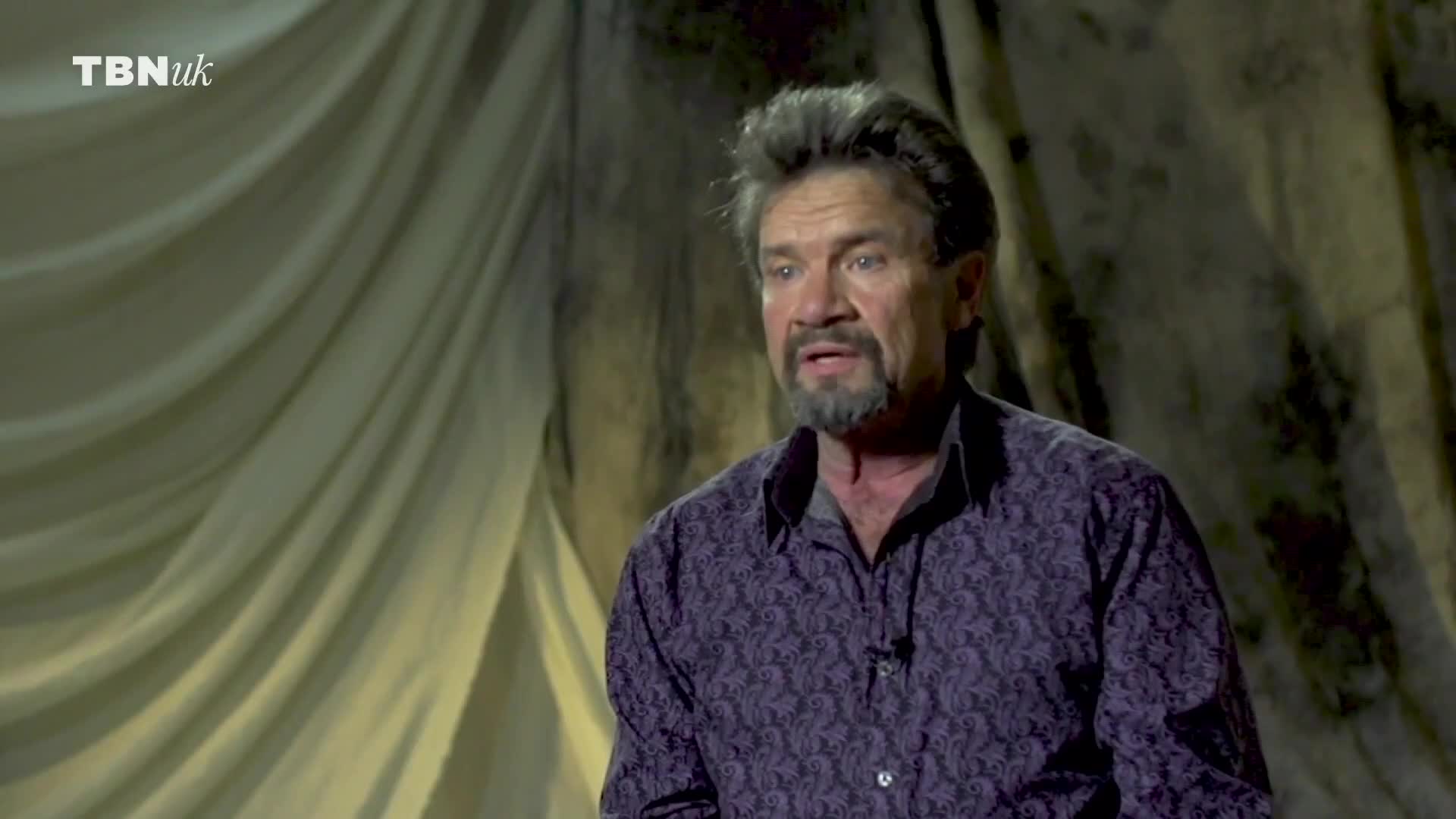 A man with a graying beard and a patterned shirt is speaking, his expression serious. Behind him, draped fabric creates a textured backdrop, and the TBN UK logo is visible.
A man with a graying beard and a patterned shirt is speaking, his expression serious. Behind him, draped fabric creates a textured backdrop, and the TBN UK logo is visible.