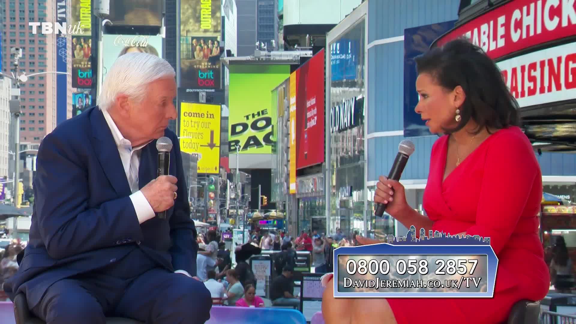 A woman in a red dress speaks into a microphone, facing a man in a suit. Behind them, bright billboards and signs fill the frame, likely in a busy public space.
A woman in a red dress speaks into a microphone, facing a man in a suit. Behind them, bright billboards and signs fill the frame, likely in a busy public space.