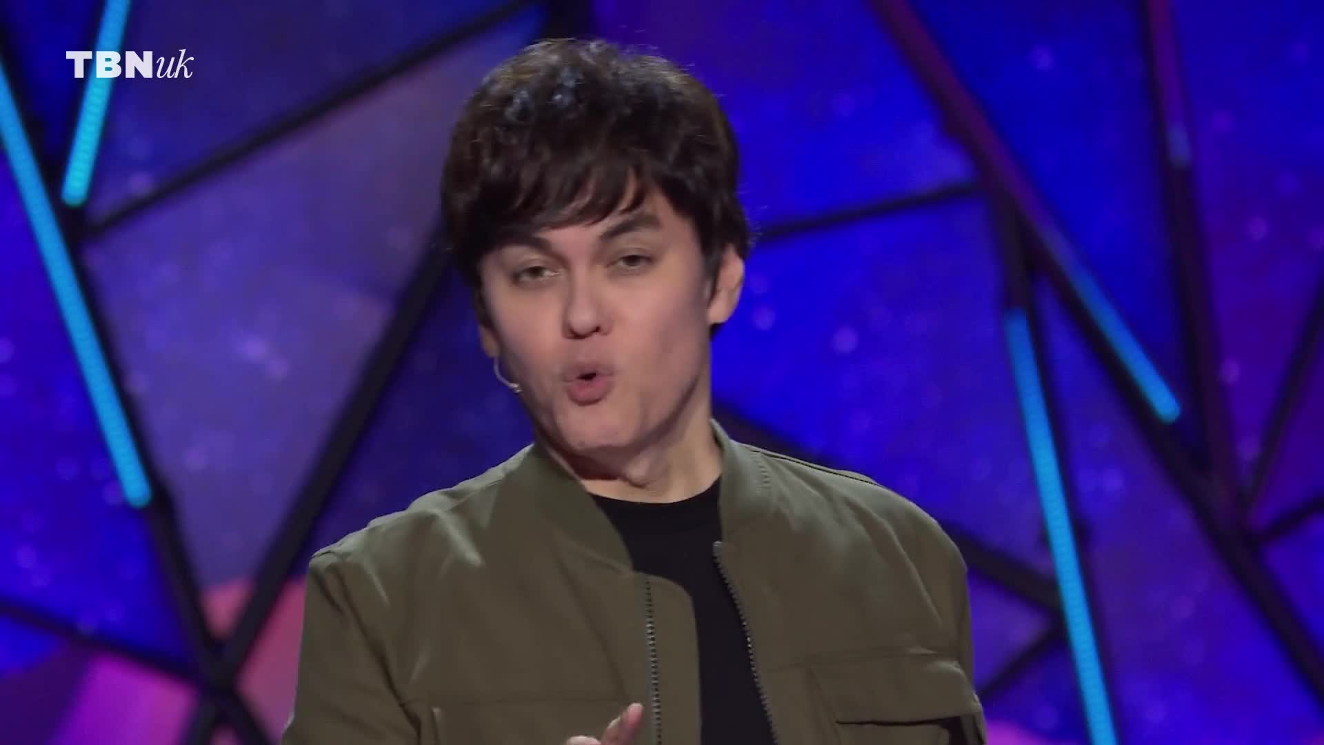 The man in the olive bomber jacket is speaking, his mouth open in a slight "O" shape. Behind him, the TBN UK logo sits against a backdrop of geometric shapes and blue light.
The man in the olive bomber jacket is speaking, his mouth open in a slight "O" shape. Behind him, the TBN UK logo sits against a backdrop of geometric shapes and blue light.