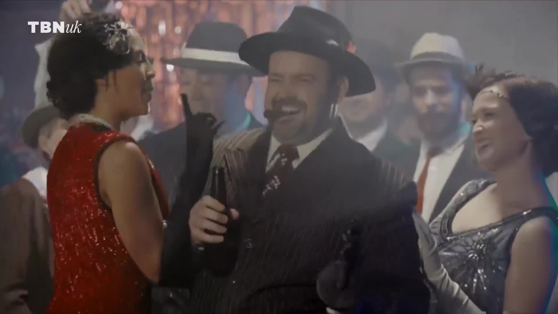 A man in a pinstripe suit and fedora grins, holding a bottle and a pipe. Around him, people in vintage attire chat and gesture in what looks like a UK-based production for TBN UK.
A man in a pinstripe suit and fedora grins, holding a bottle and a pipe. Around him, people in vintage attire chat and gesture in what looks like a UK-based production for TBN UK.