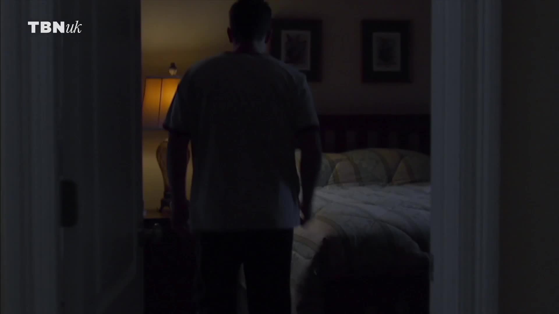 A man in a t-shirt walks toward a lit bedroom, the doorway framing his silhouette. The warm glow of a bedside lamp illuminates the bed and the room beyond.
A man in a t-shirt walks toward a lit bedroom, the doorway framing his silhouette. The warm glow of a bedside lamp illuminates the bed and the room beyond.