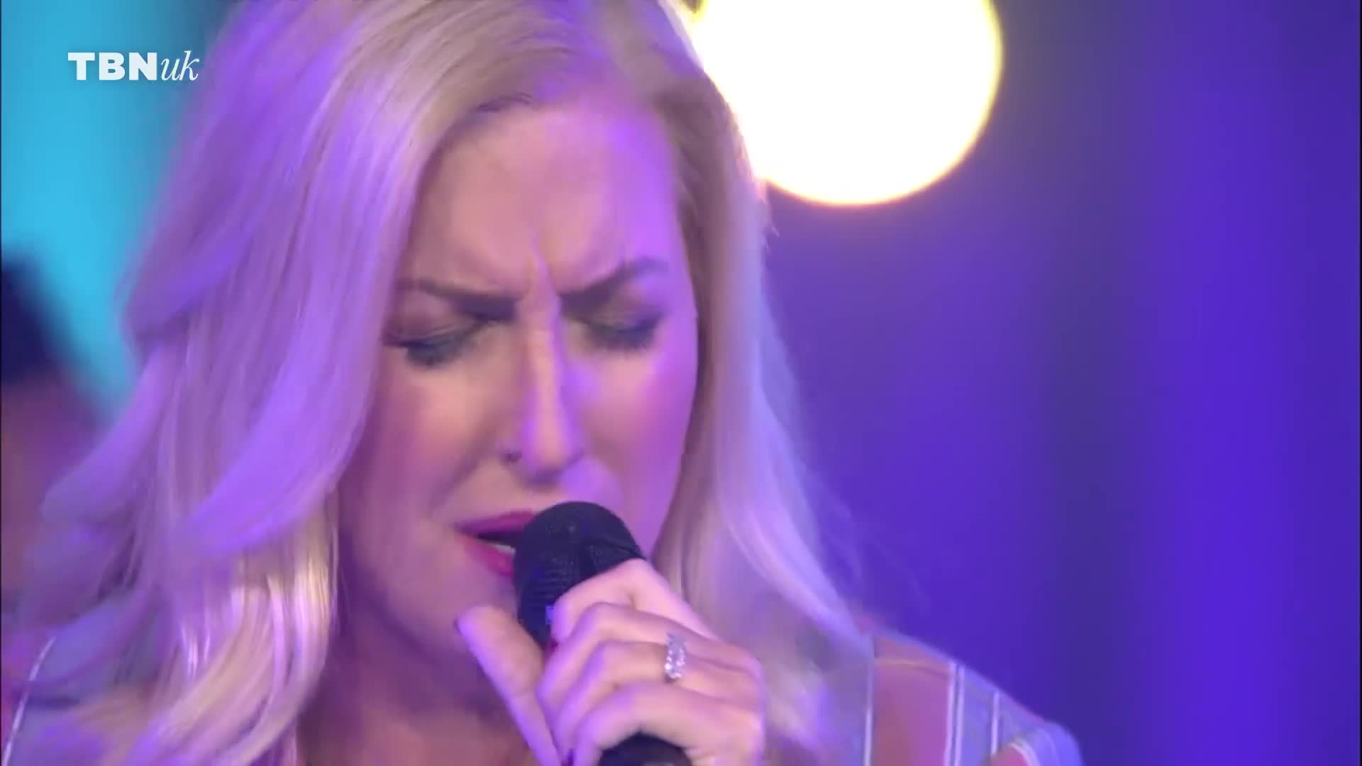 The woman is singing into a microphone, her face contorted with emotion. Above her, the TBN UK logo is visible.
The woman is singing into a microphone, her face contorted with emotion. Above her, the TBN UK logo is visible.