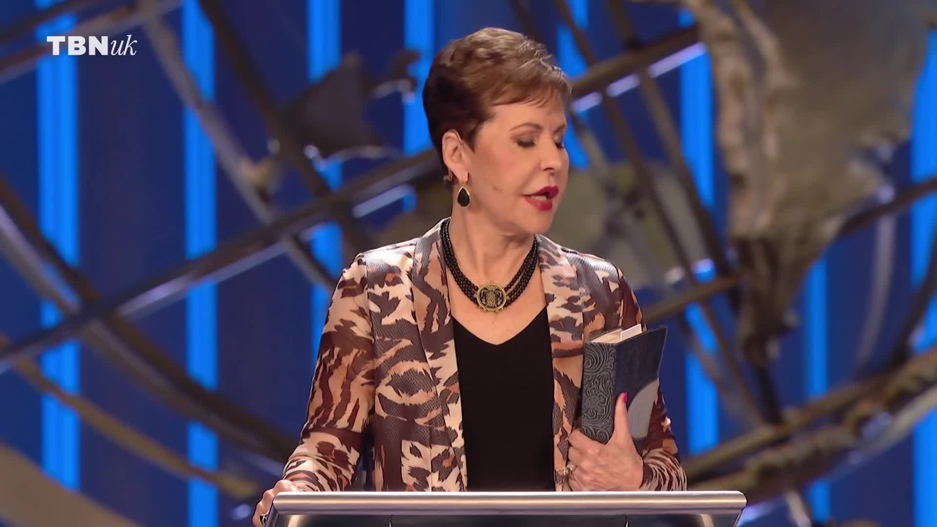 A woman in a patterned jacket stands behind a lectern, holding a book. The TBN UK logo is visible in the upper left corner.