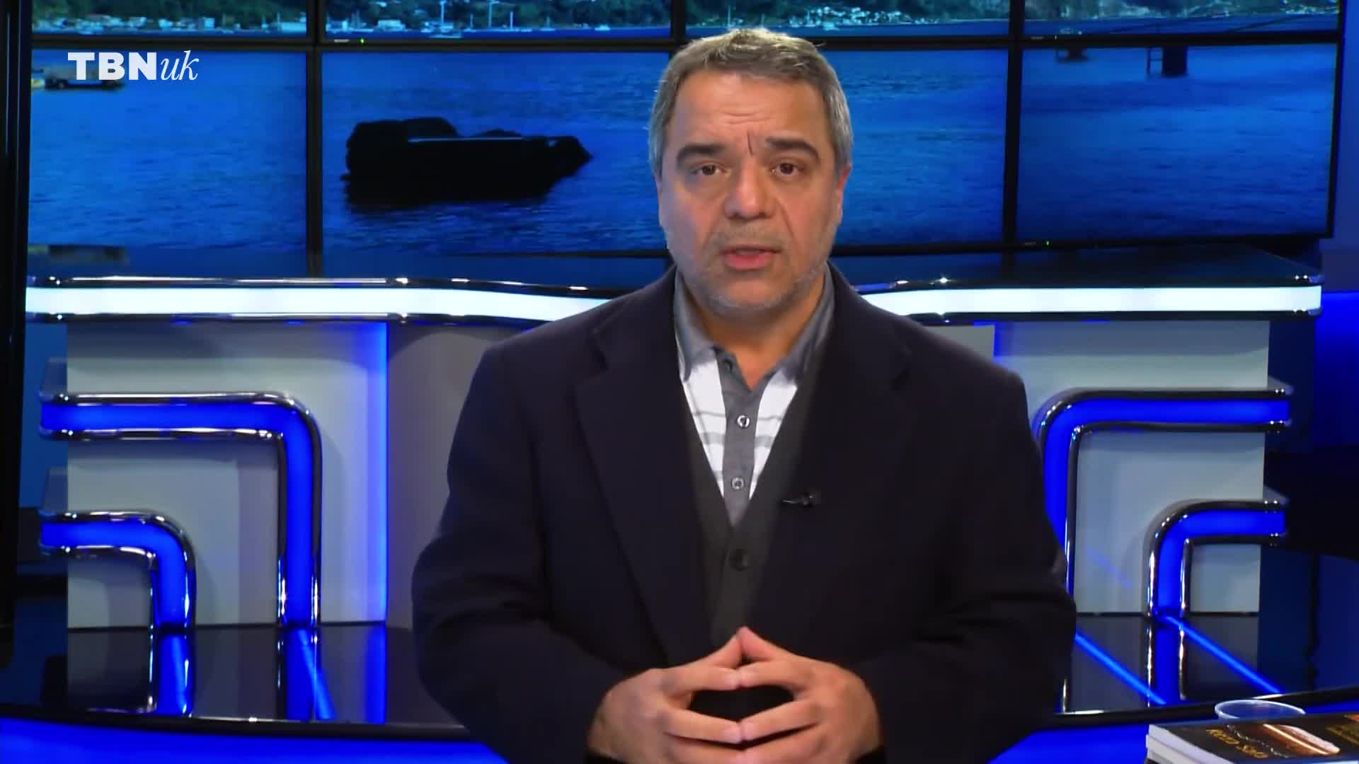 A man stands before a studio backdrop showing a harbour scene, his hands clasped. The TBN UK logo is visible in the upper left corner.