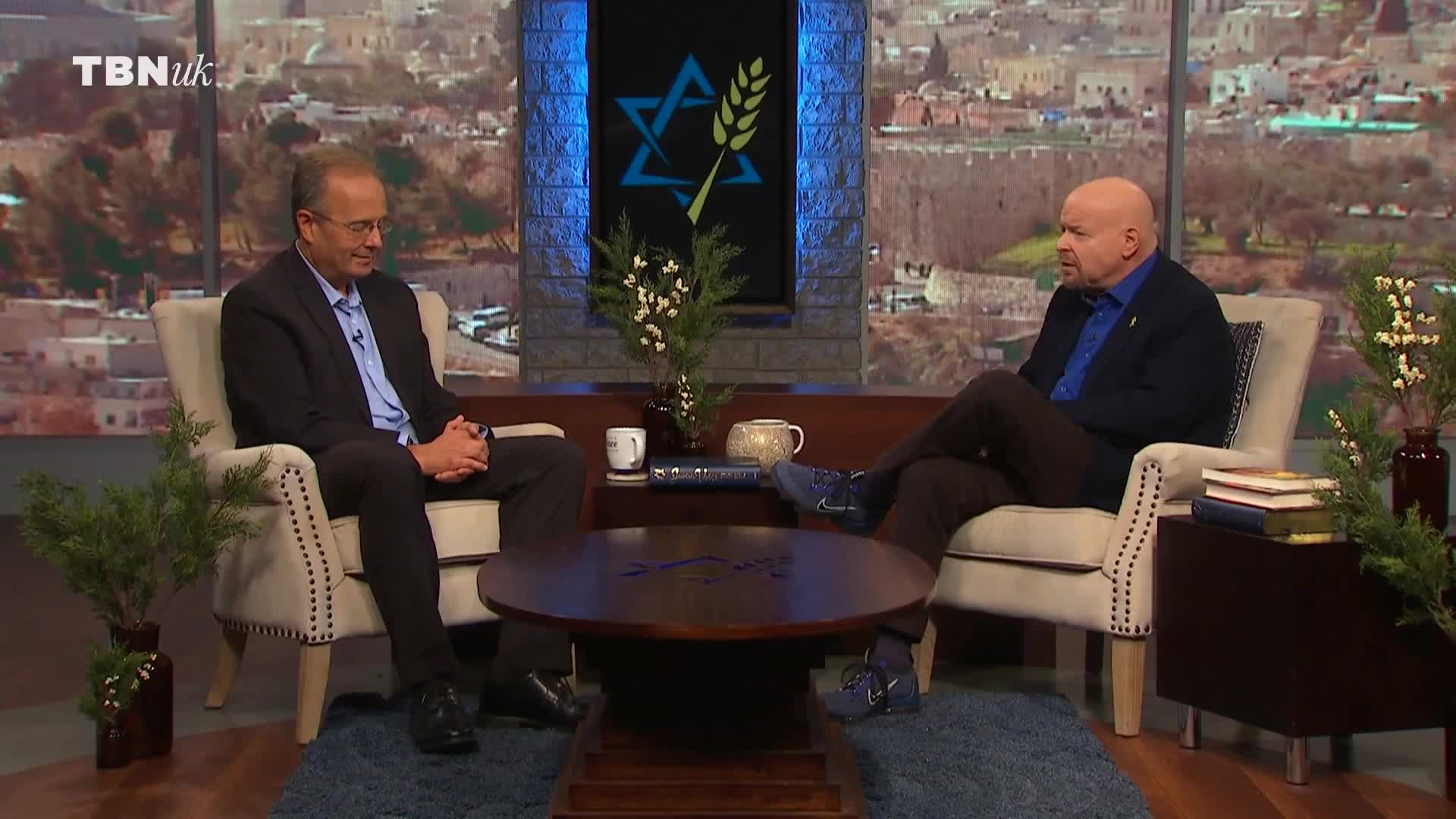 Two men sit in plush armchairs, facing each other across a dark wood coffee table. A TBN UK logo is visible in the corner of the screen.