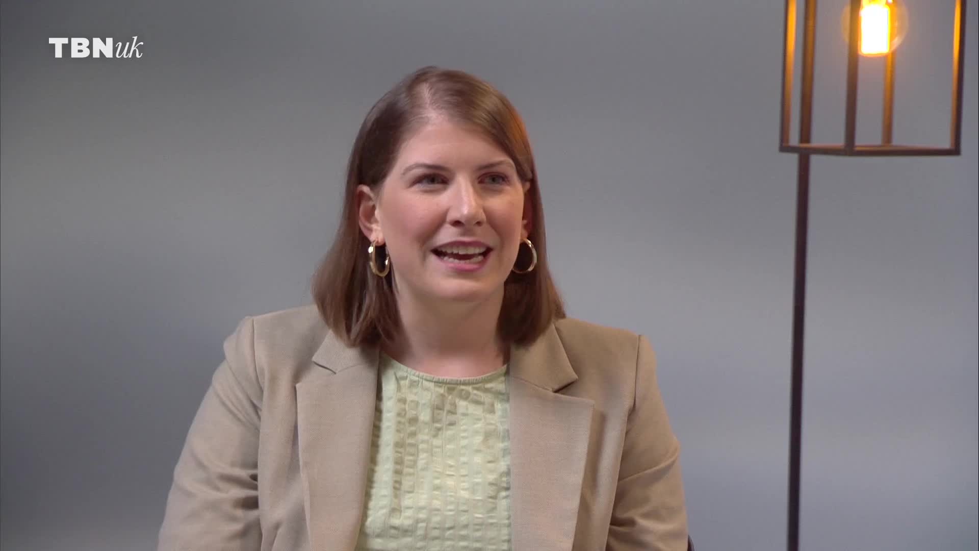 A woman in a tan blazer and a light green patterned top speaks directly to the camera. A tall, modern floor lamp with a single bulb stands to her right.