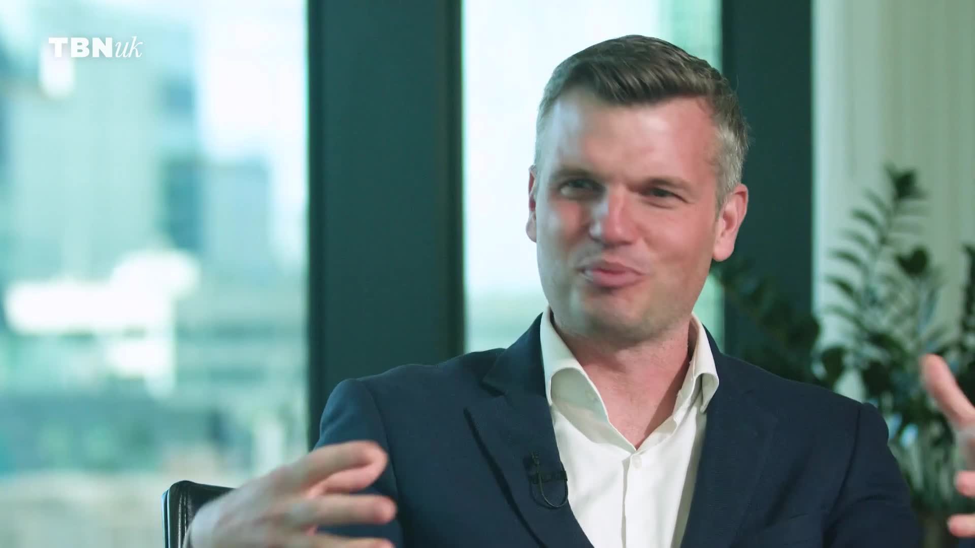 A man in a dark suit and white shirt speaks animatedly, gesturing with his hands. The TBN UK logo is visible in the top left corner.