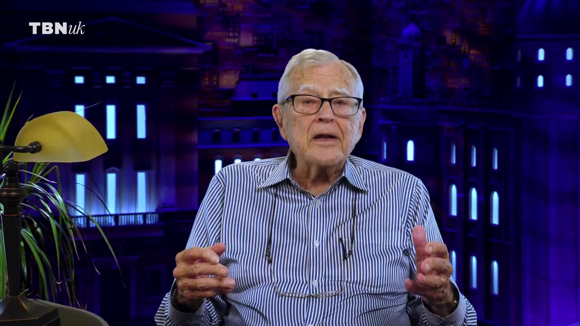 An older man with glasses speaks, his hands gesturing. Behind him, a backdrop depicts a building with illuminated windows. The TBN UK logo appears in the corner.