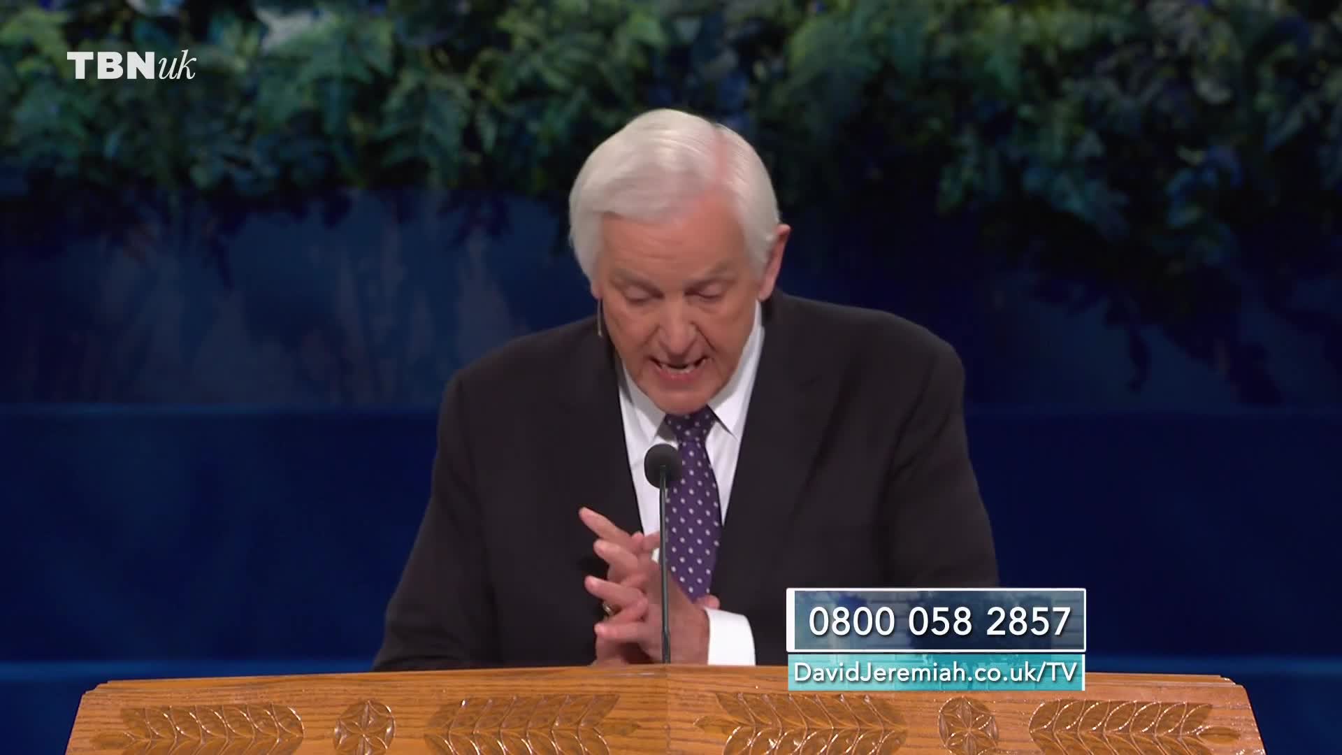 A man in a dark suit and patterned tie speaks into a microphone, his hands clasped. A phone number and website are displayed on a graphic in the lower right corner of the screen, alongside the TBN UK logo.