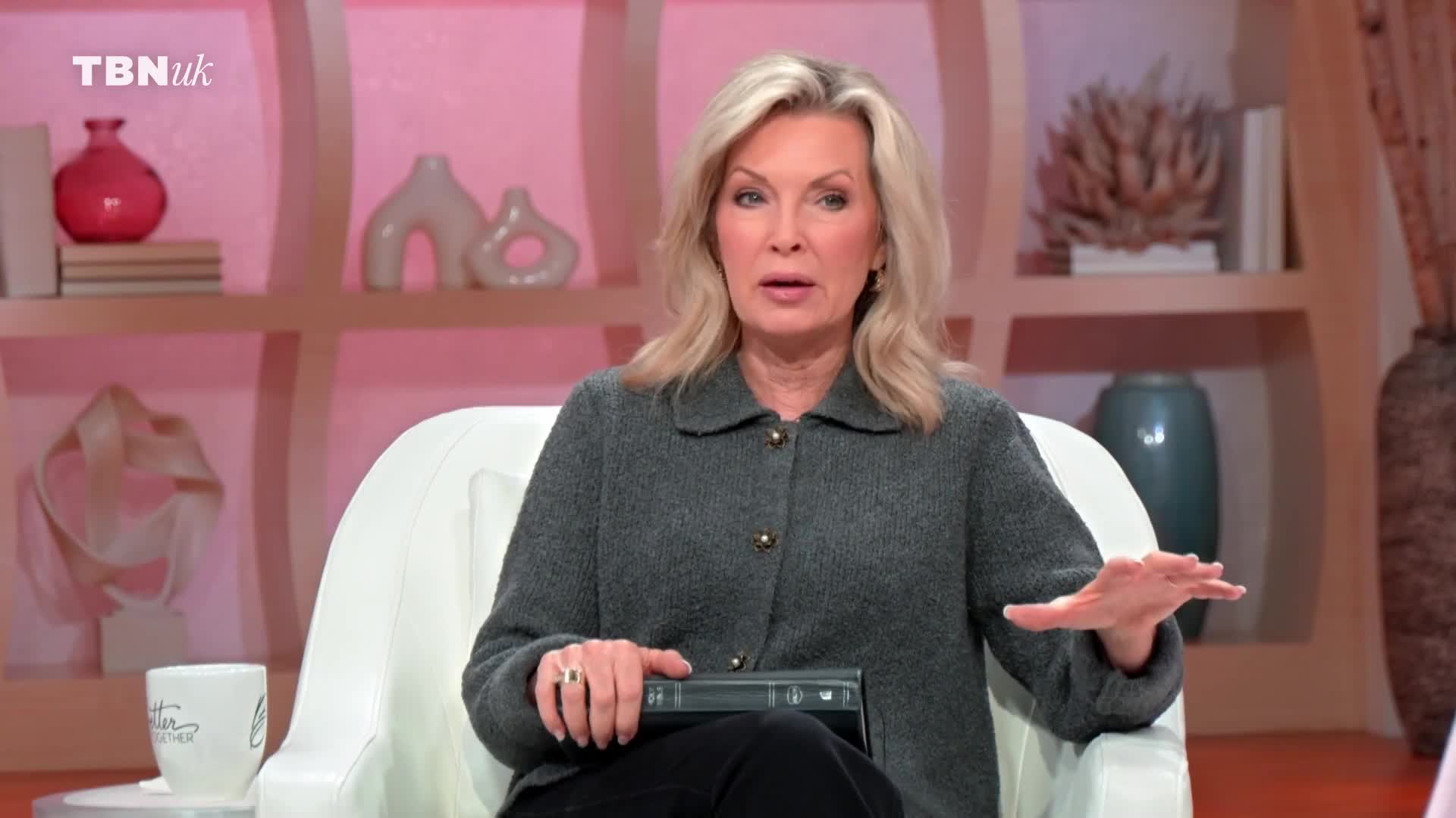 A woman with blonde hair is speaking, her hand gesturing outwards. She's seated on a white sofa, a thick book resting on her lap.
