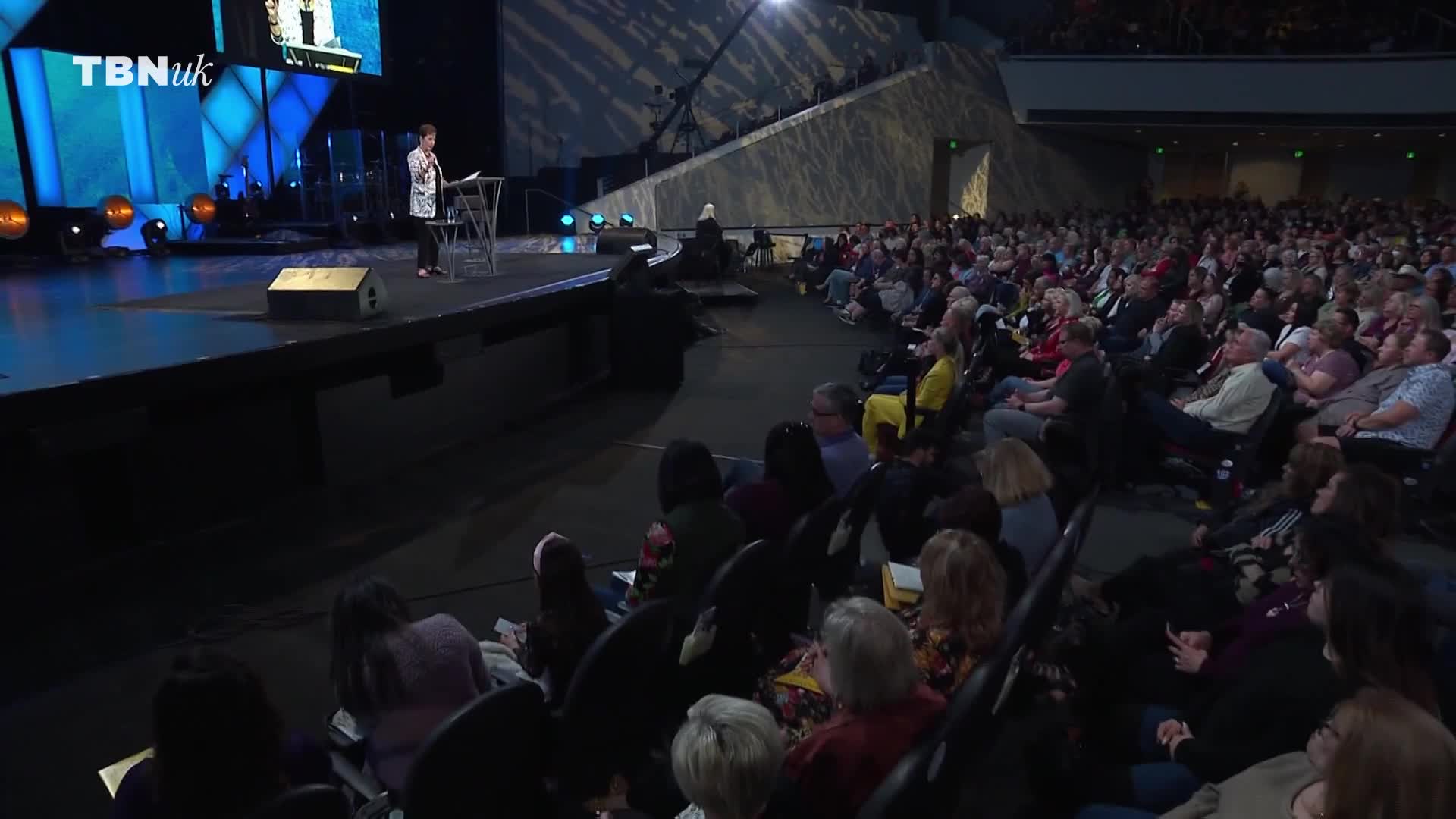 A woman stands at a podium on a large stage, addressing a packed auditorium. The TBN UK logo is visible on a screen behind her.