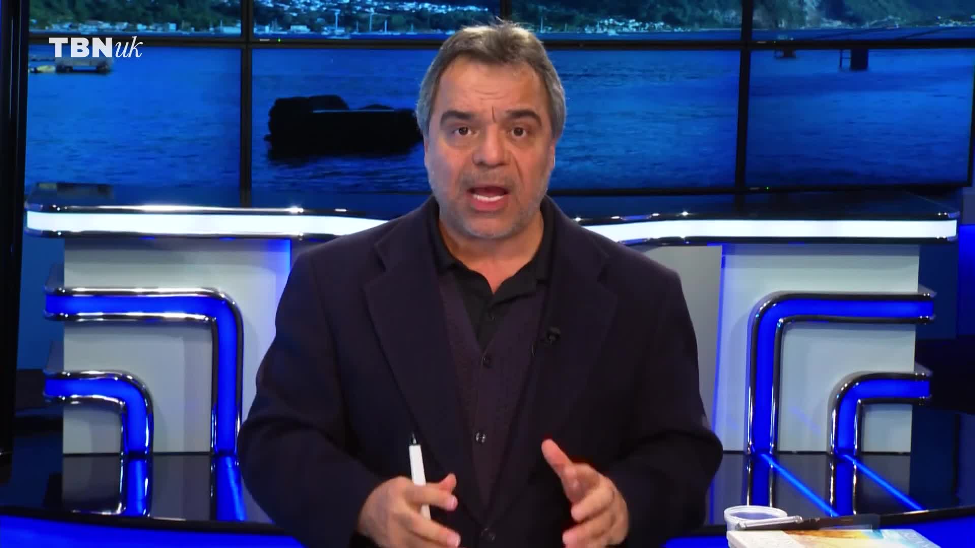 A man in a dark suit stands behind a desk with blue lighting. He gestures with a pen in his right hand, speaking directly to the camera. The backdrop behind him displays a serene seascape with a TBN UK logo in the corner.