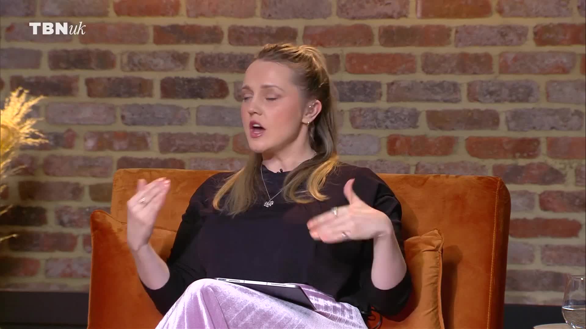 A woman with her hair in a ponytail speaks animatedly, gesturing with her hands. She sits on an orange chair in front of a brick wall, with the TBN UK logo visible.
