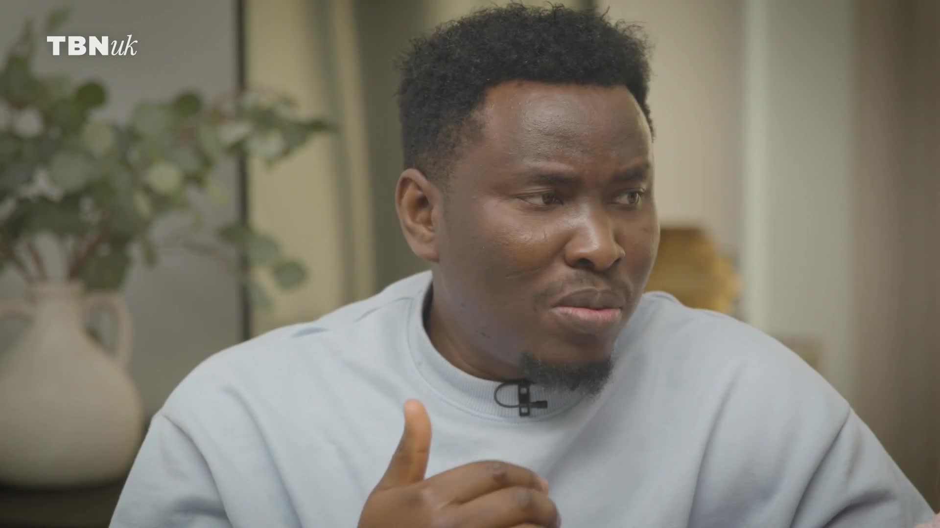 A man with dark, curly hair gestures with his right hand while speaking. The TBN UK logo appears in the upper left corner of the frame.