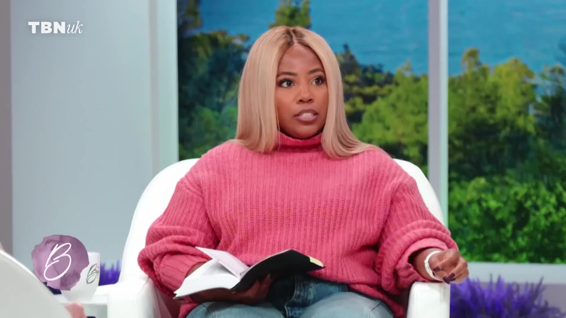 A woman with blonde hair and a bright pink sweater is speaking, holding an open book in her lap. The TBN UK logo is visible in the upper left corner.