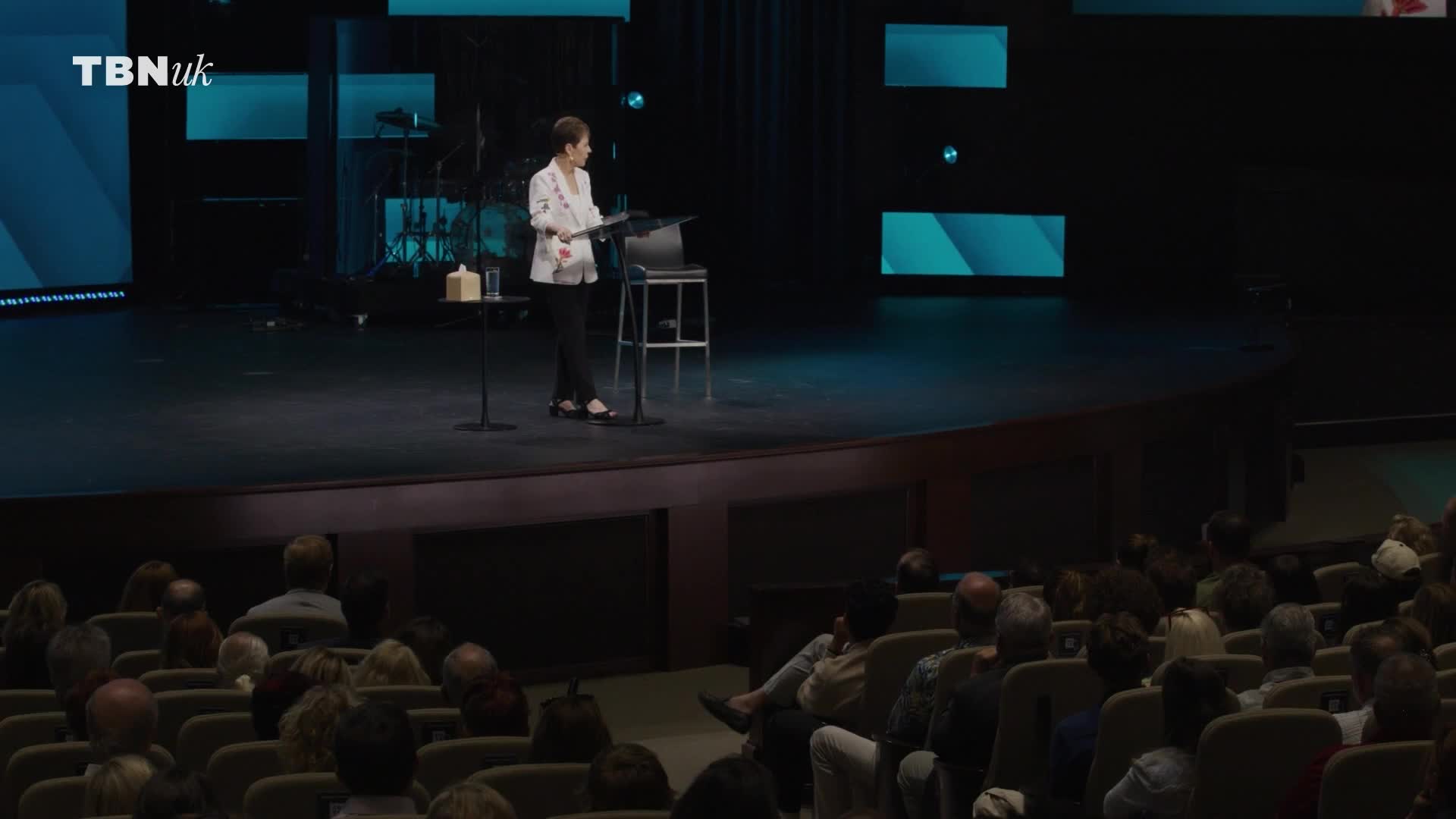 A woman stands at a lectern on a stage, addressing an audience. The TBN UK logo is visible in the top left corner.