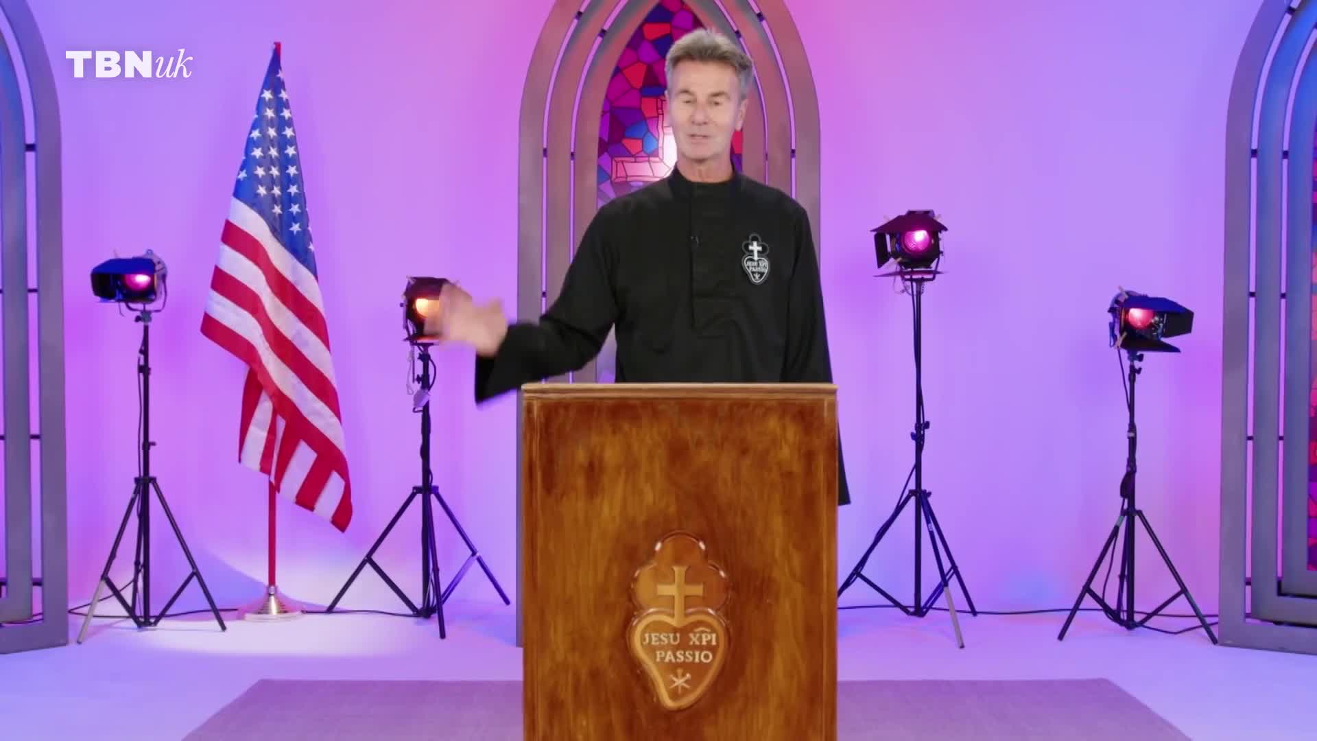 A man in a black clerical shirt stands at a wooden lectern, gesturing with his left hand. An American flag stands to his left, illuminated by stage lights.