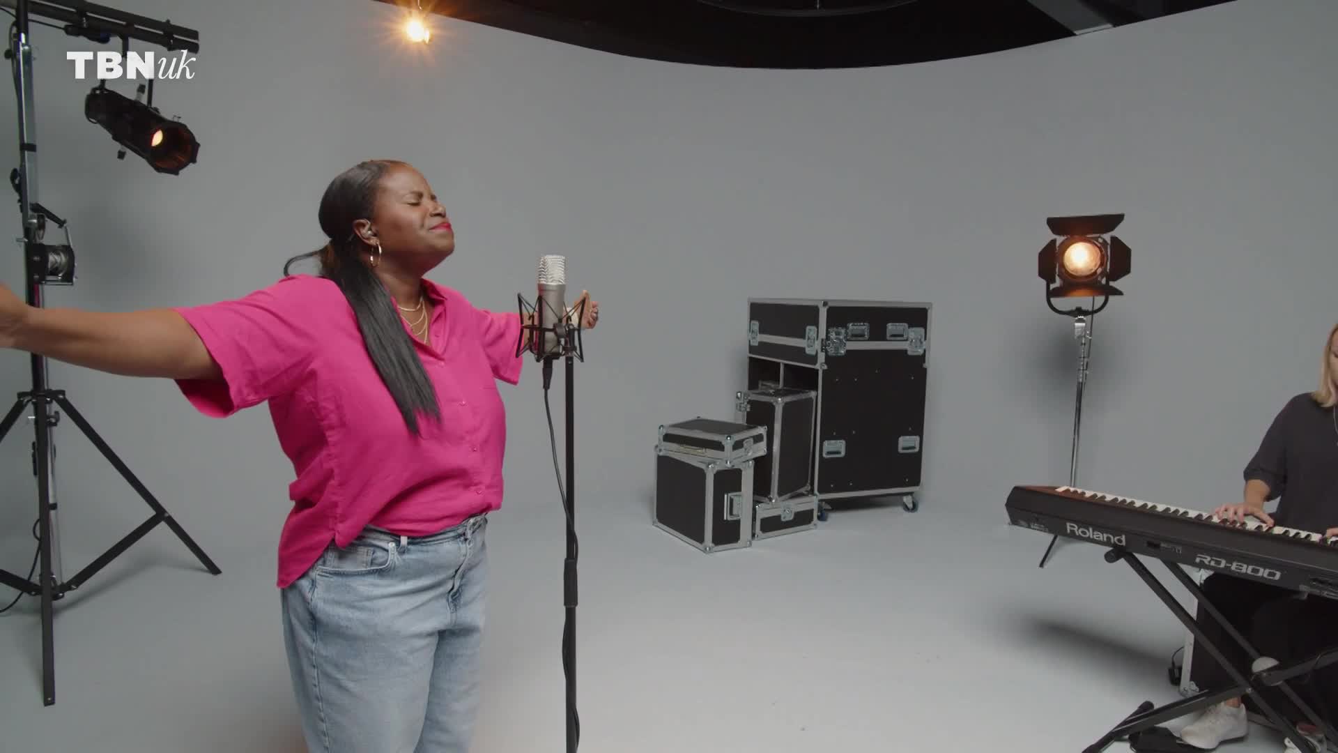 A woman in a bright pink shirt belts into a microphone, her eyes closed in passionate song. Beside her, another woman plays a Roland keyboard, her fingers moving across the keys.