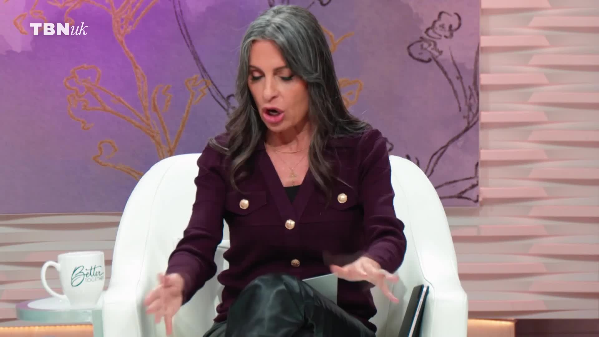 The woman on TBN UK gestures emphatically as she speaks, her hands moving to emphasize her words. A white mug with "Better" written on it sits on a side table beside her.