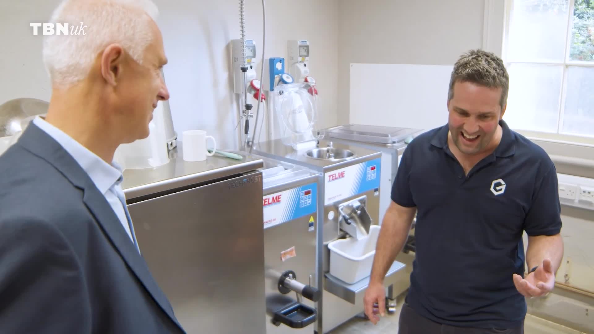 A man in a suit turns to speak with another man in a dark polo shirt. The second man laughs, gesturing with his hands near some stainless steel equipment.