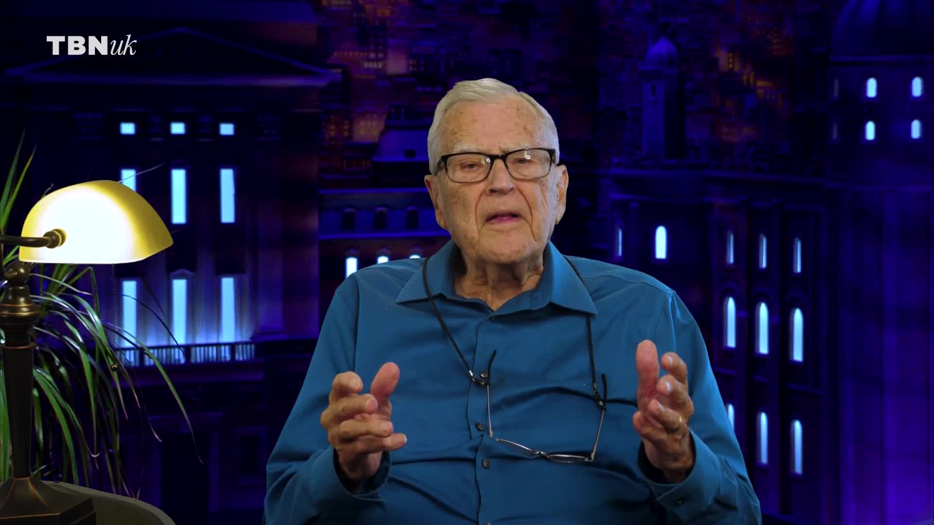 A man in a blue shirt speaks with his hands gesturing. Behind him, a TBN UK logo appears over a backdrop of illuminated buildings.