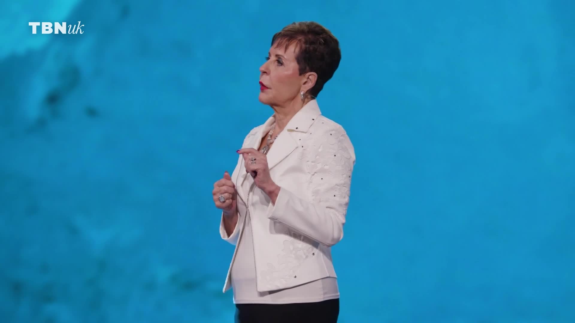 A woman in a white jacket stands against a bright blue backdrop, gesturing with her hands. The TBN UK logo appears in the upper left corner.