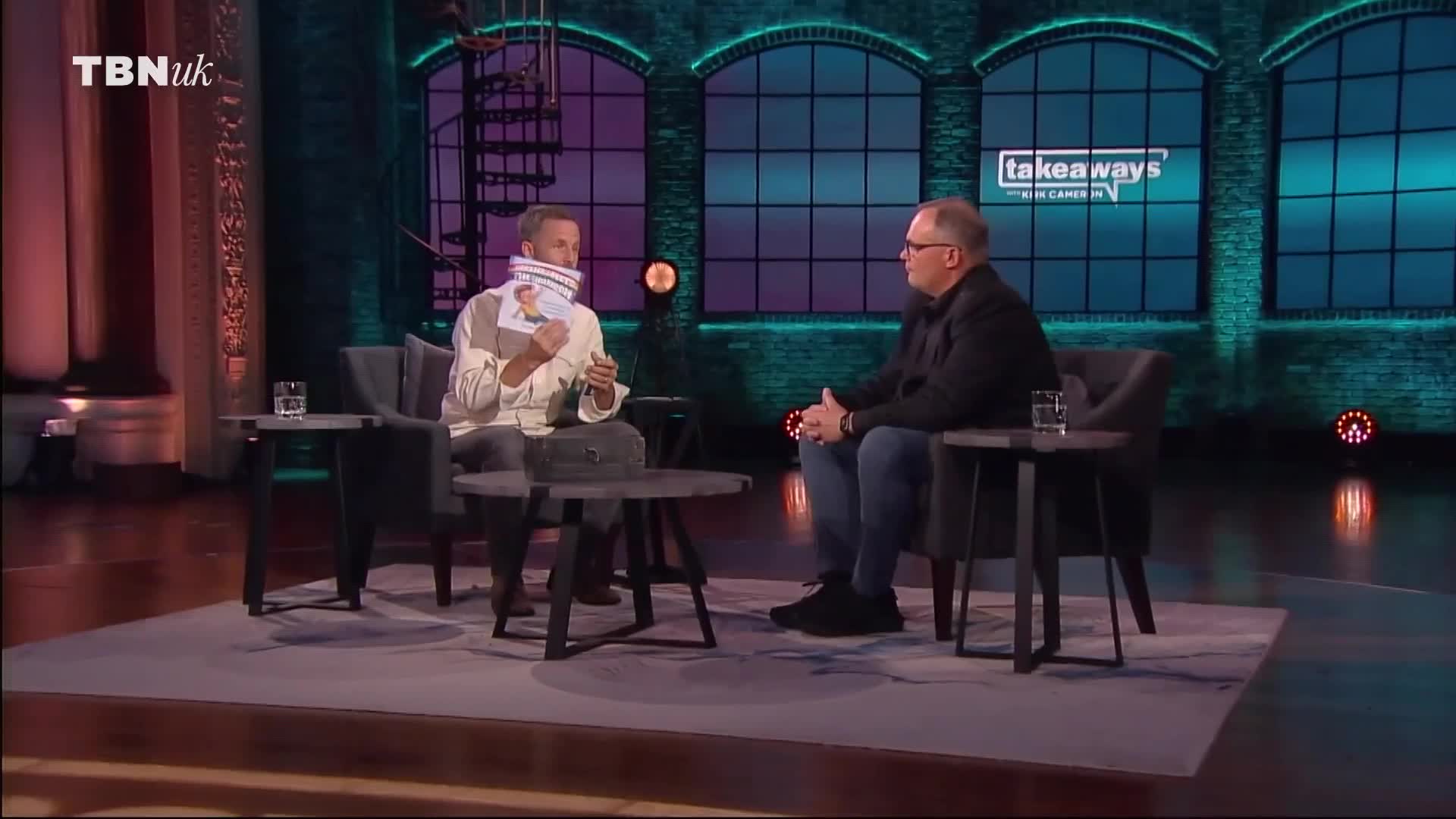Two men are seated in armchairs on a stage, facing each other. The man on the left holds up a pamphlet, while the man on the right listens intently. The TBN UK logo is visible in the upper left corner.