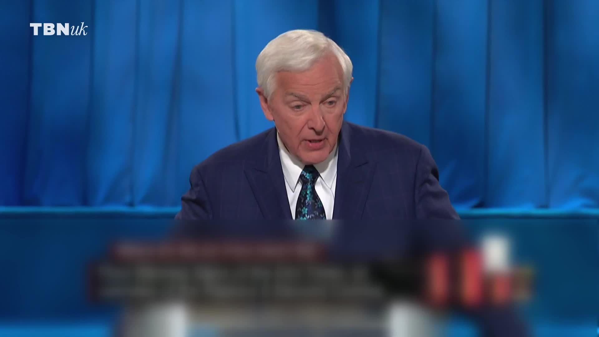 A man in a blue suit and patterned tie speaks animatedly. The TBN UK logo appears in the top left corner.